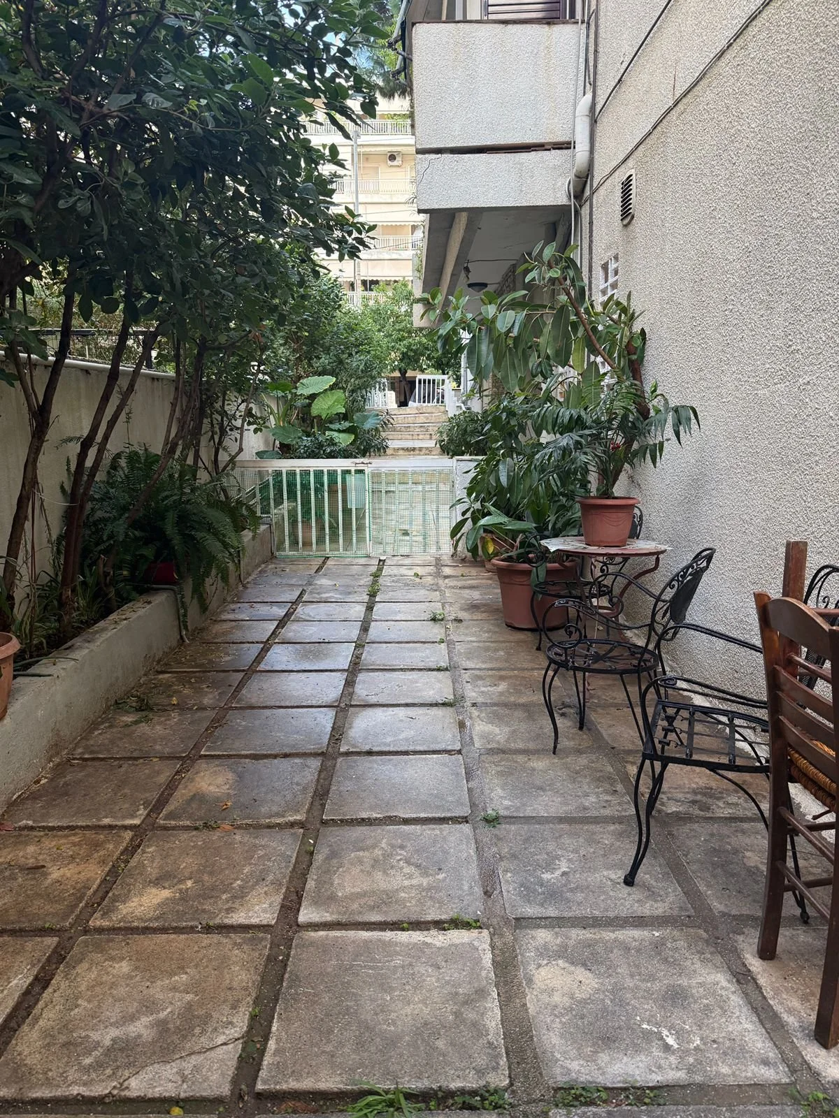 A small outdoor patio area with potted plants, trees, and outdoor furniture, enclosed by a low wall and gated at the end, with a concrete pavement surface.