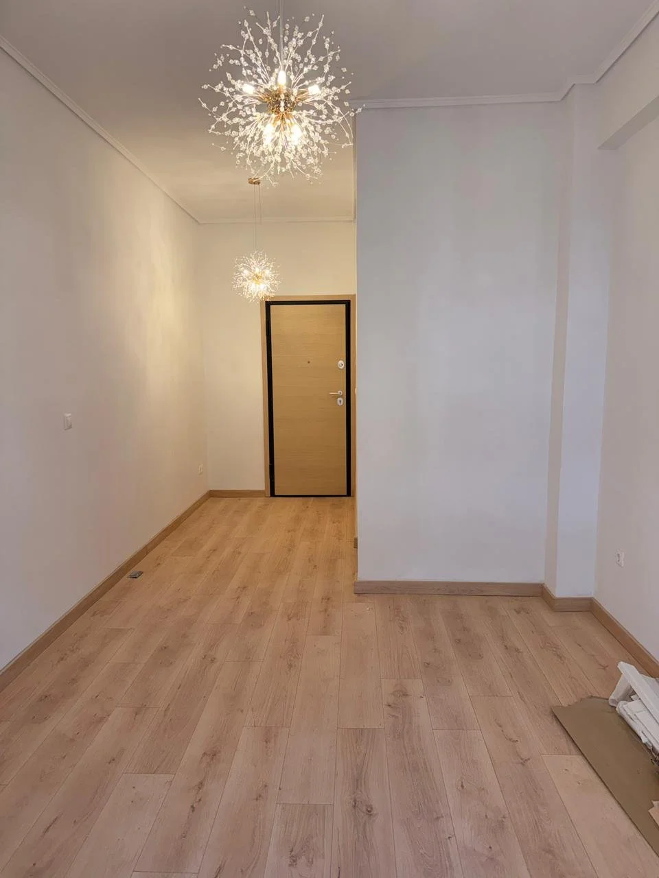 Empty hallway with light-colored wooden flooring, white walls, a wooden door at the end, and modern chandelier ceiling lights.