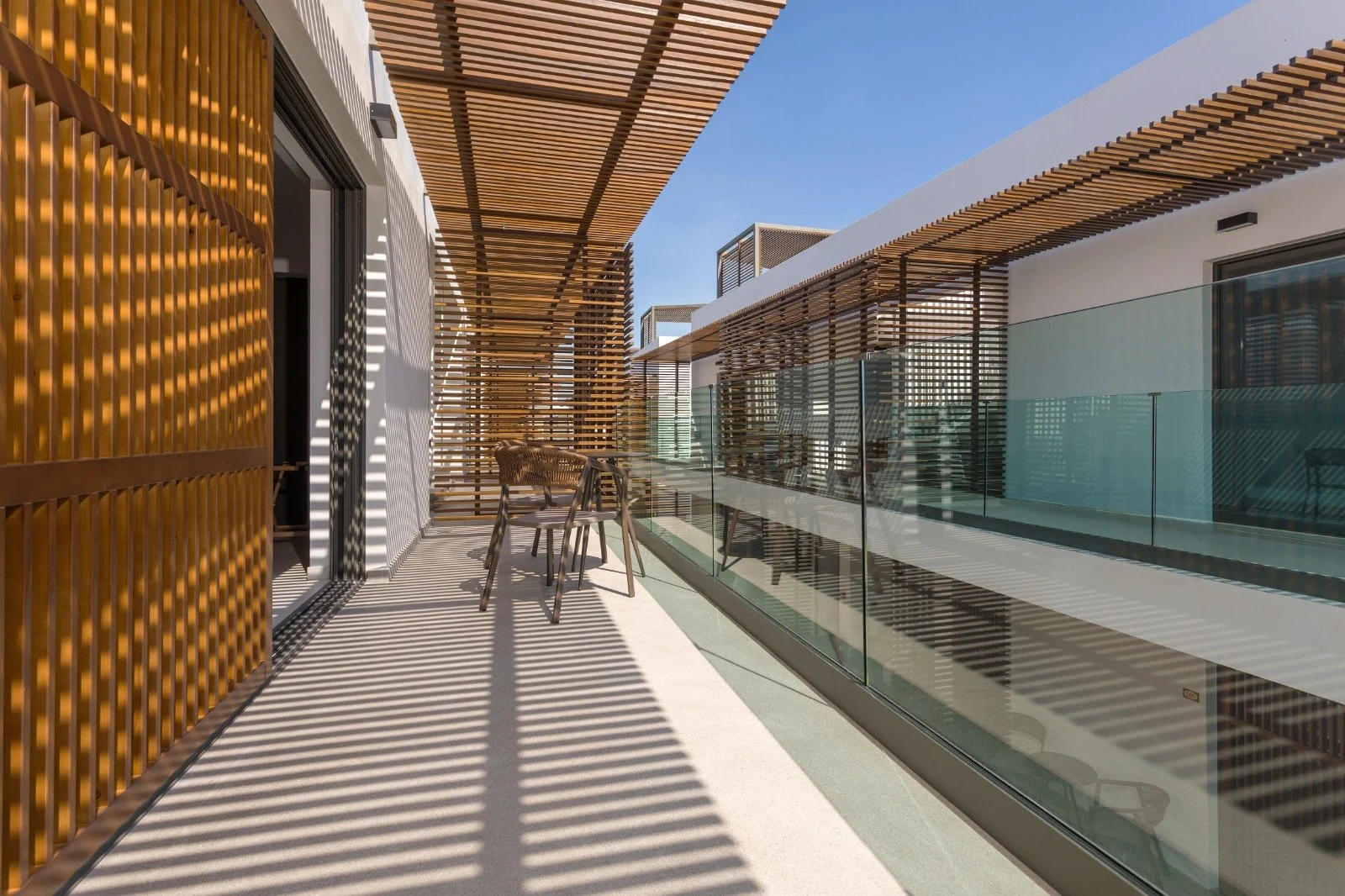 Modern balcony with wooden slats creating striped shadows, metal and glass railing, and outdoor furniture, under a clear blue sky.