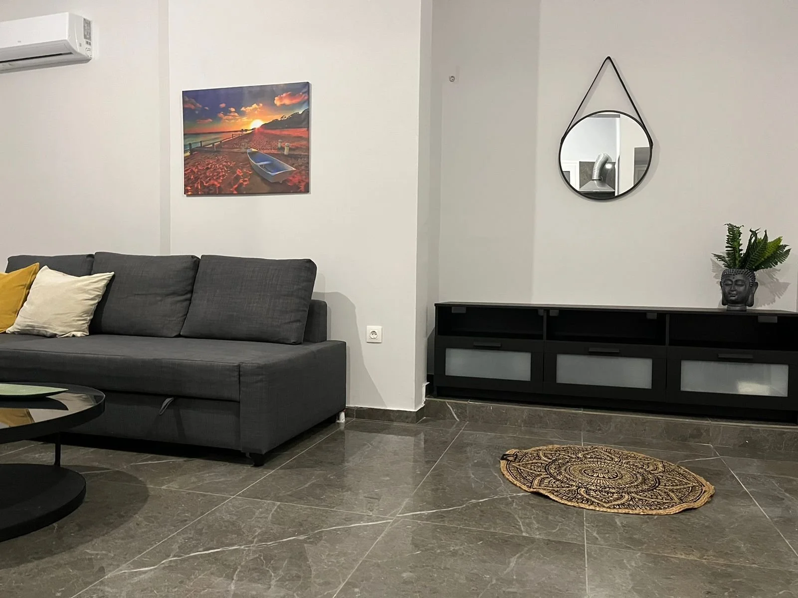 Living room with a gray sofa, beige and yellow pillows, a black coffee table, a black sideboard, a decorative rug, wall art of a sunset and boat, a mirror, a potted plant, and an air conditioning unit.