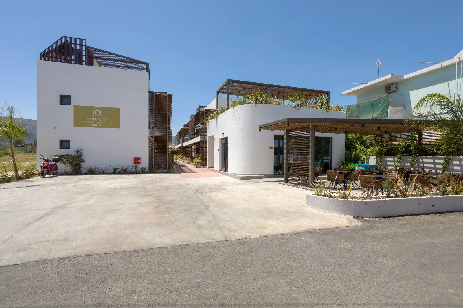 Exterior view of a modern hotel named Serenity located in Agruja, with white building facades, outdoor seating area with wooden chairs, and a motorcycle parked in front under a clear blue sky.