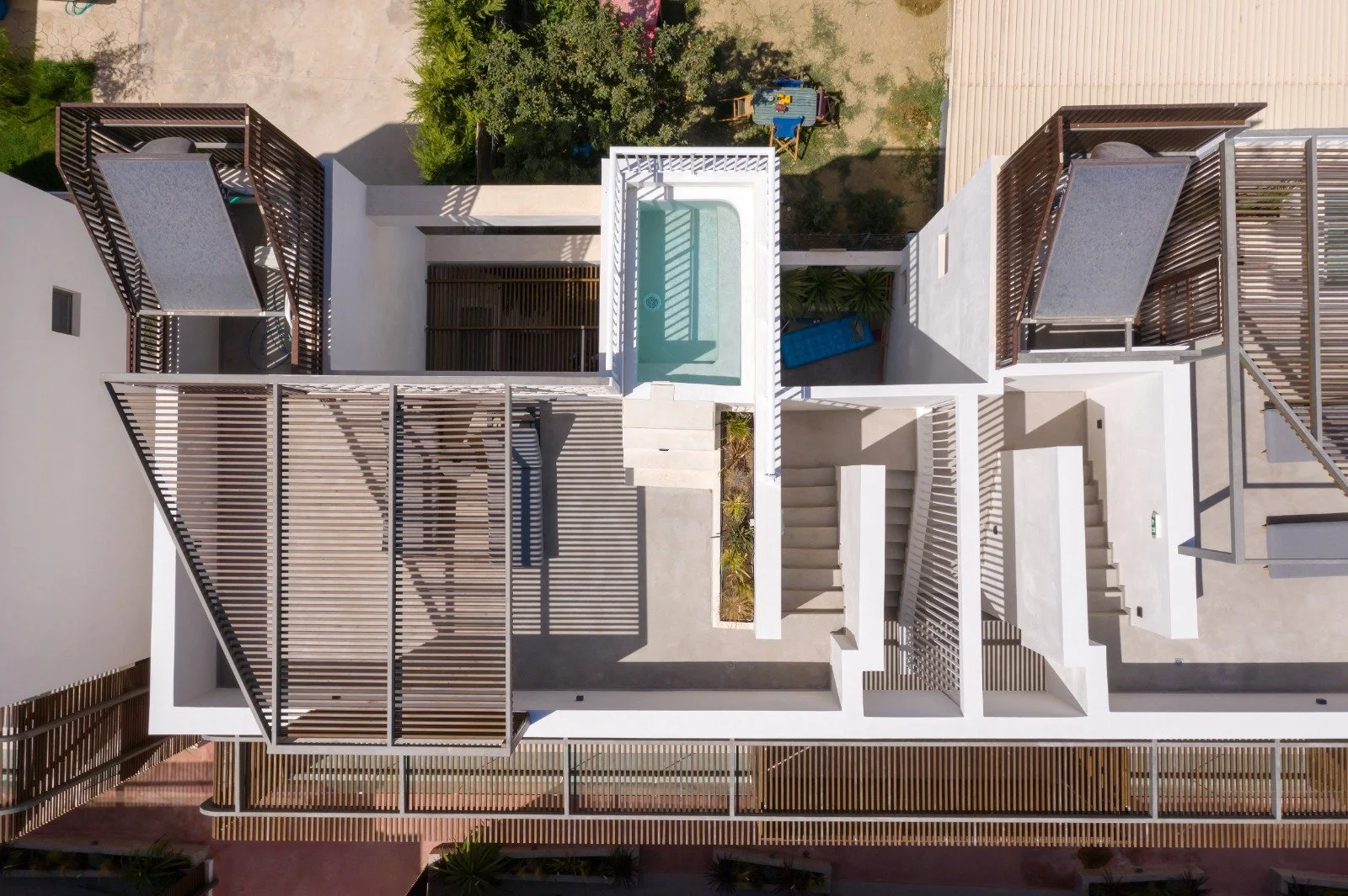 An aerial view of a modern building's rooftop with shaded terraces, a small pool, and outdoor furniture, surrounded by trees and outdoor areas.