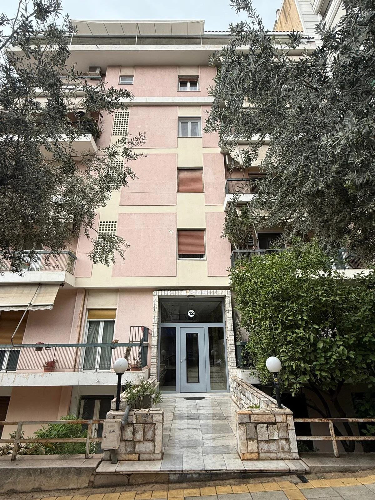Front view of a multi-story pink residential building with balconies and windows, entryway with glass door, surrounded by trees and plants, with street and sidewalk in foreground. Greece, Investment, Golden Visa, Real Estate, Cyprus, Georgia, Lebanon