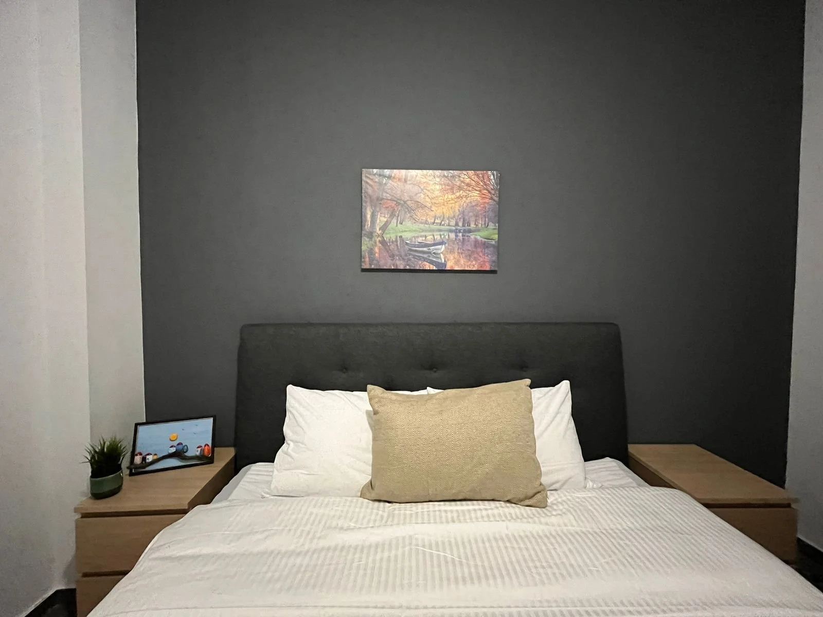 A neatly made bed with a dark upholstered headboard, two white pillows, and a beige decorative pillow, flanked by two wooden side tables. The left side table holds a small potted plant and a tablet device displaying a cartoon. Above the bed, there's 