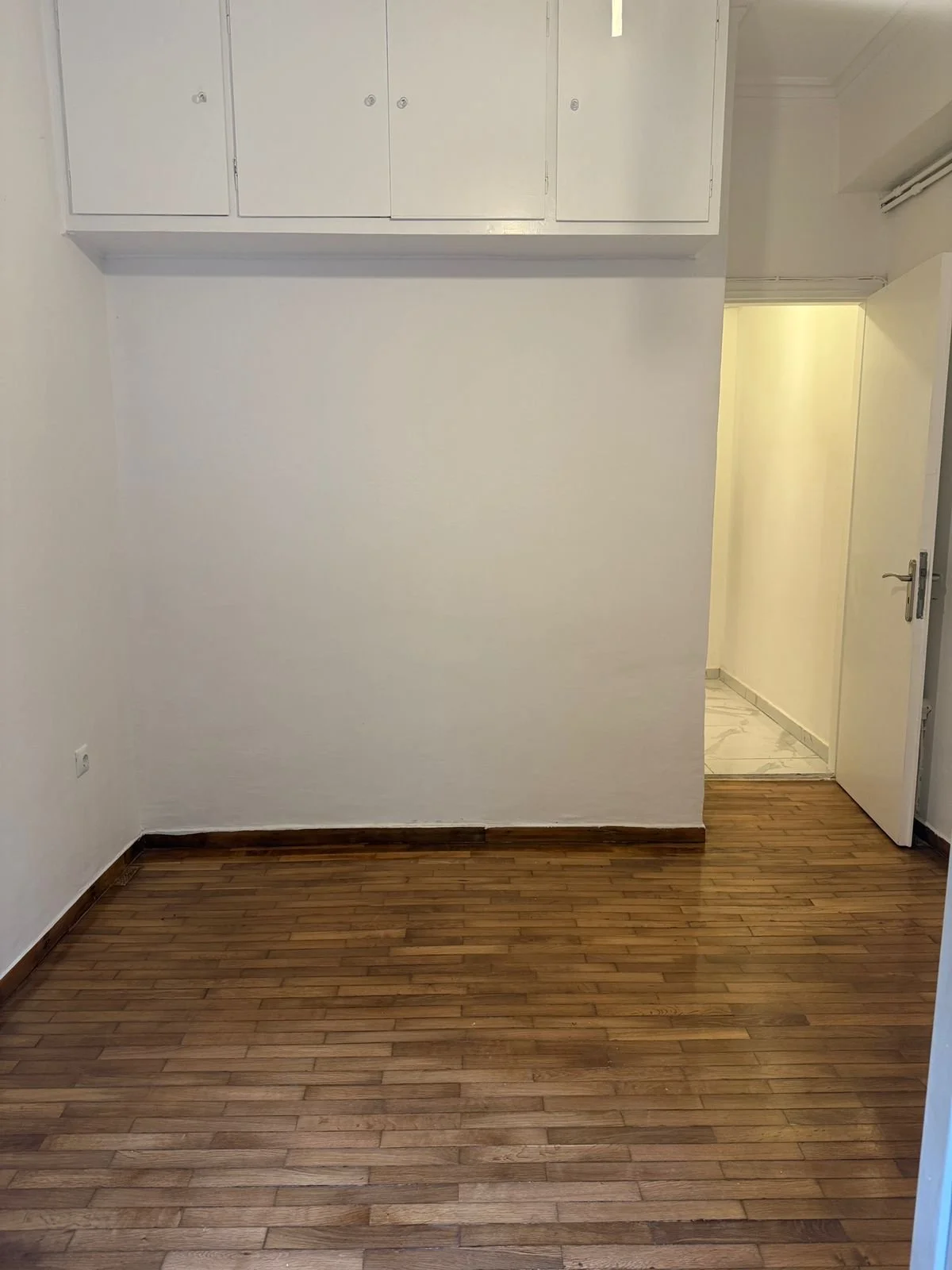 Empty room with wooden floor, white wall, ceiling cabinets, door open to a hallway with marble floor.