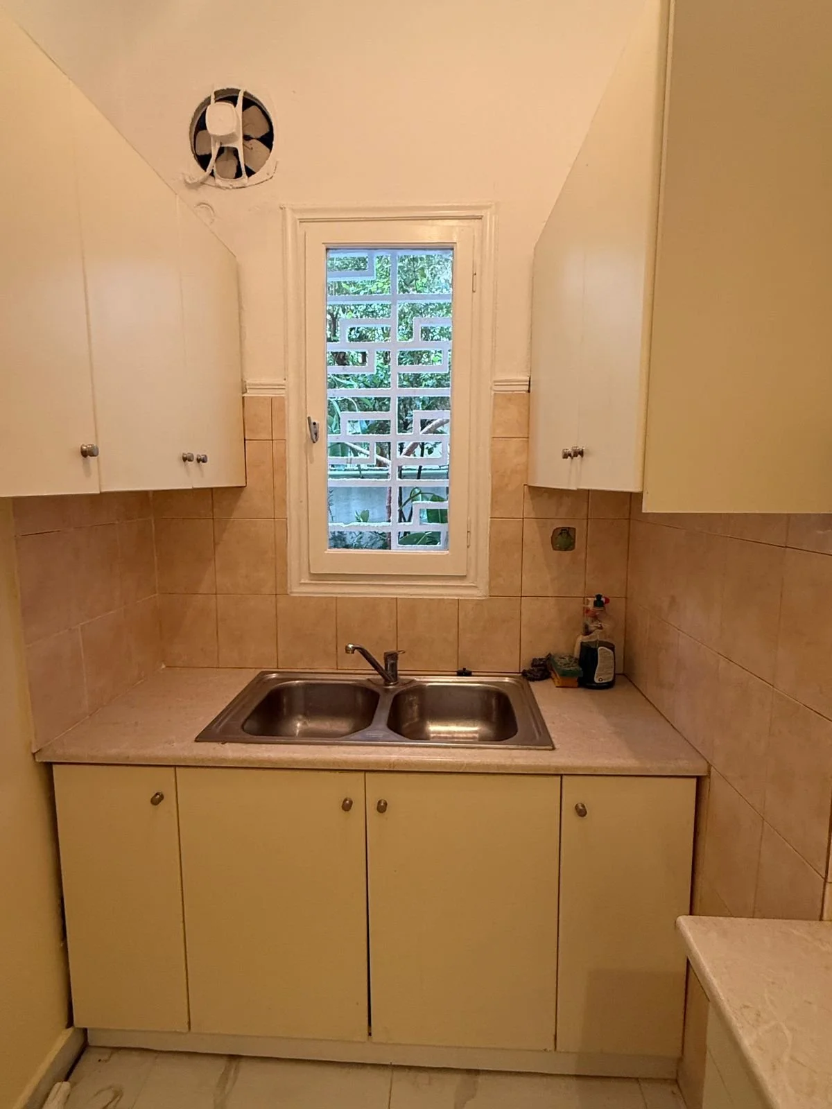 Small kitchen with a double sink, beige cabinets, beige tiled backsplash, a window with a decorative grill, and a wall-mounted fan in the ceiling.