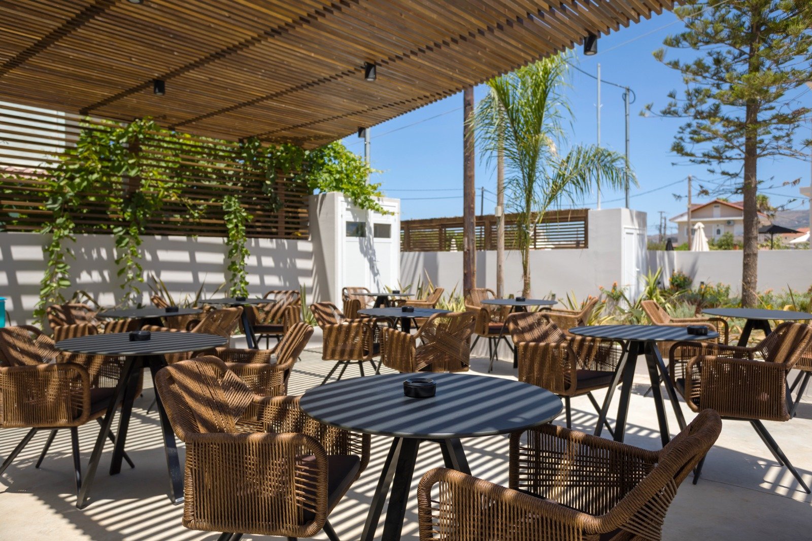 Outdoor patio with wooden tables and wicker chairs, shaded by a wooden slat roof, with palm trees and a white fence in the background under a clear blue sky.