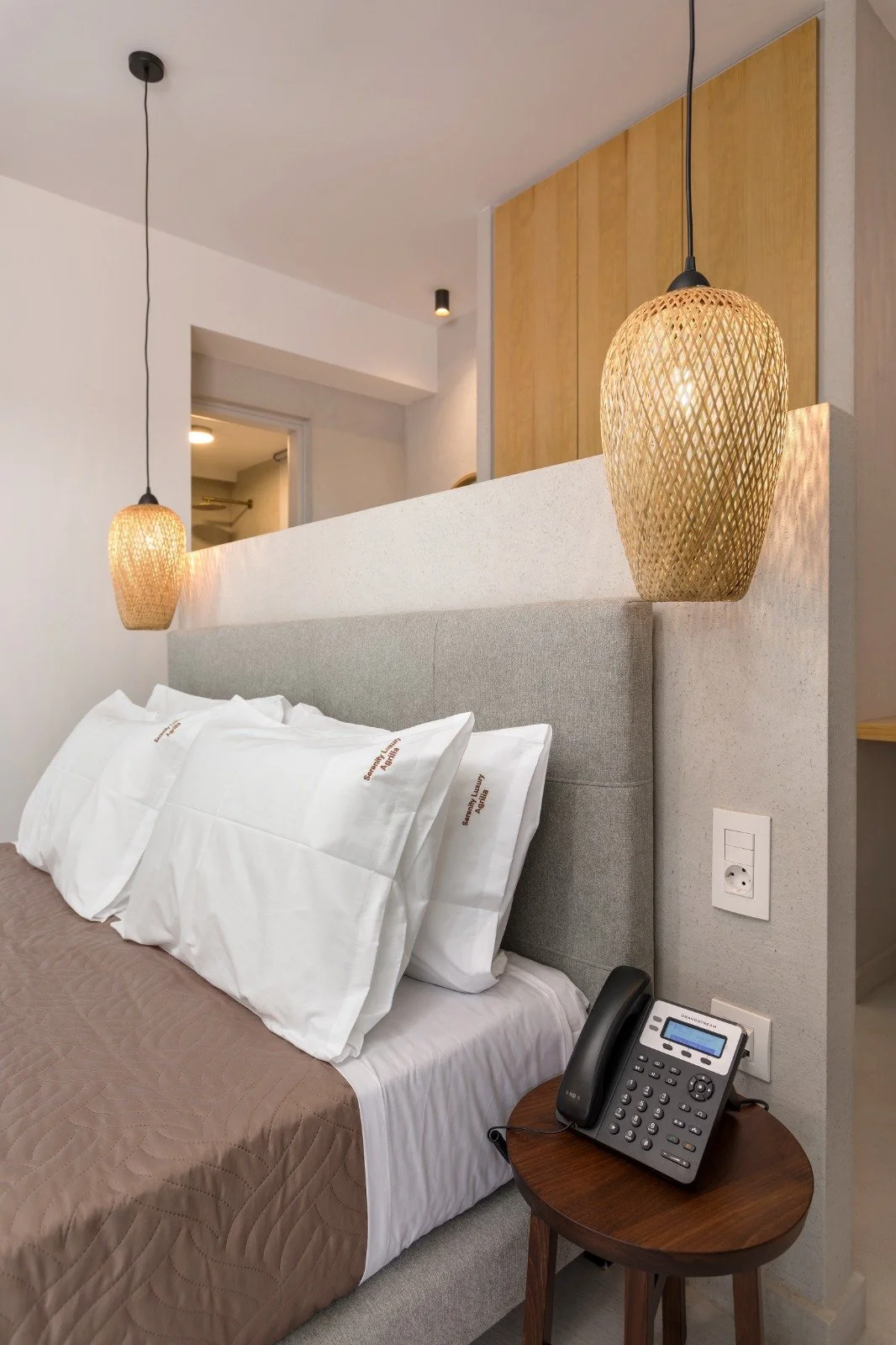 A hotel room with a bed, pillows, and bedside table with a telephone, with two woven pendant lights hanging above the bed, and a wooden panel behind the headboard.