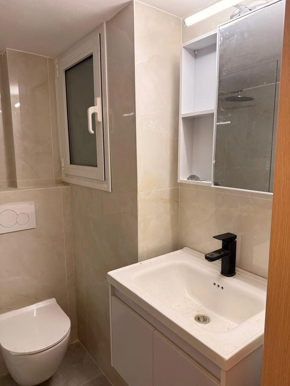 A small bathroom with beige tiled walls, a white toilet, a white sink with a black faucet, a small open medicine cabinet with shelves, a wall-mounted mirror, and a small window.