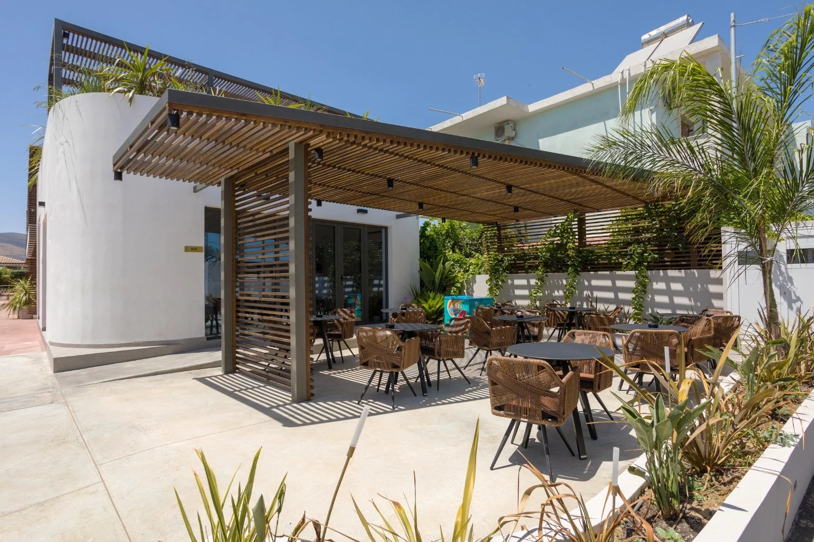 Outdoor patio area of a restaurant or café with tables and chairs under a wooden pergola, surrounded by plants and greenery, with a modern white building in the background.