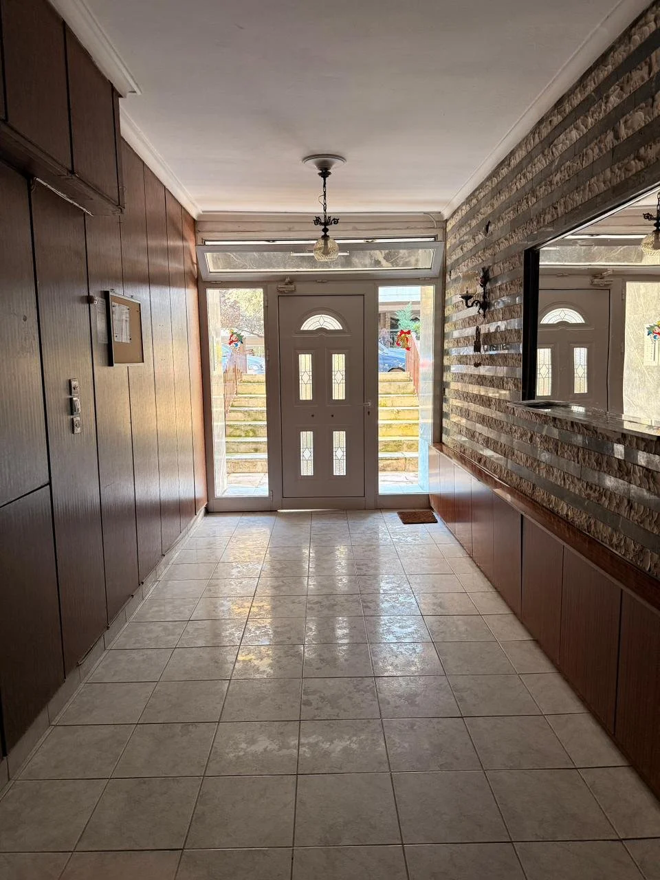 Empty entryway with tiled floor, wood-paneled walls on the left, brick accent wall on the right with a large mirror, front door with small windows, and stairs with Christmas decorations outside.