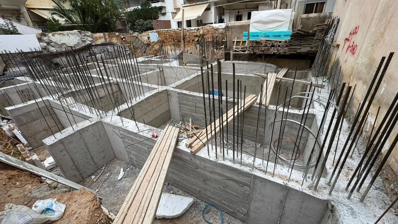 Construction site showing concrete foundation sections with rebar reinforcement, wooden planks, and construction materials, likely for building a structure.