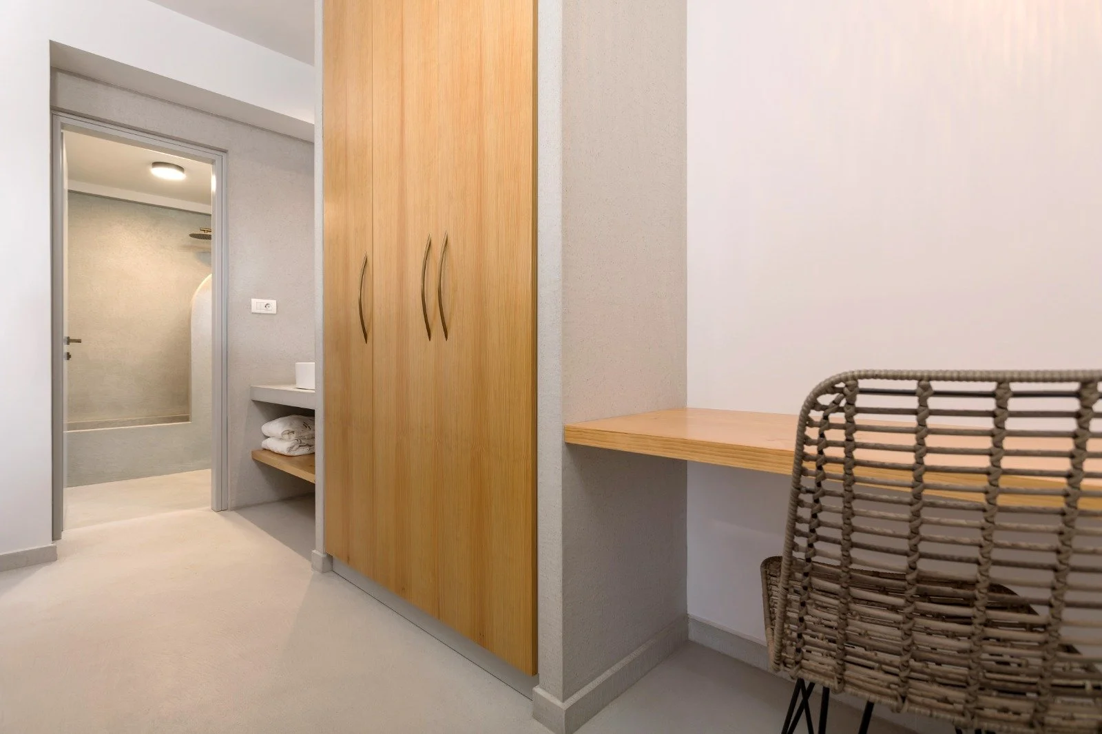 Room with a wooden wardrobe, a small desk with a woven chair, and an open bathroom door showing a shower and a linen shelf.
