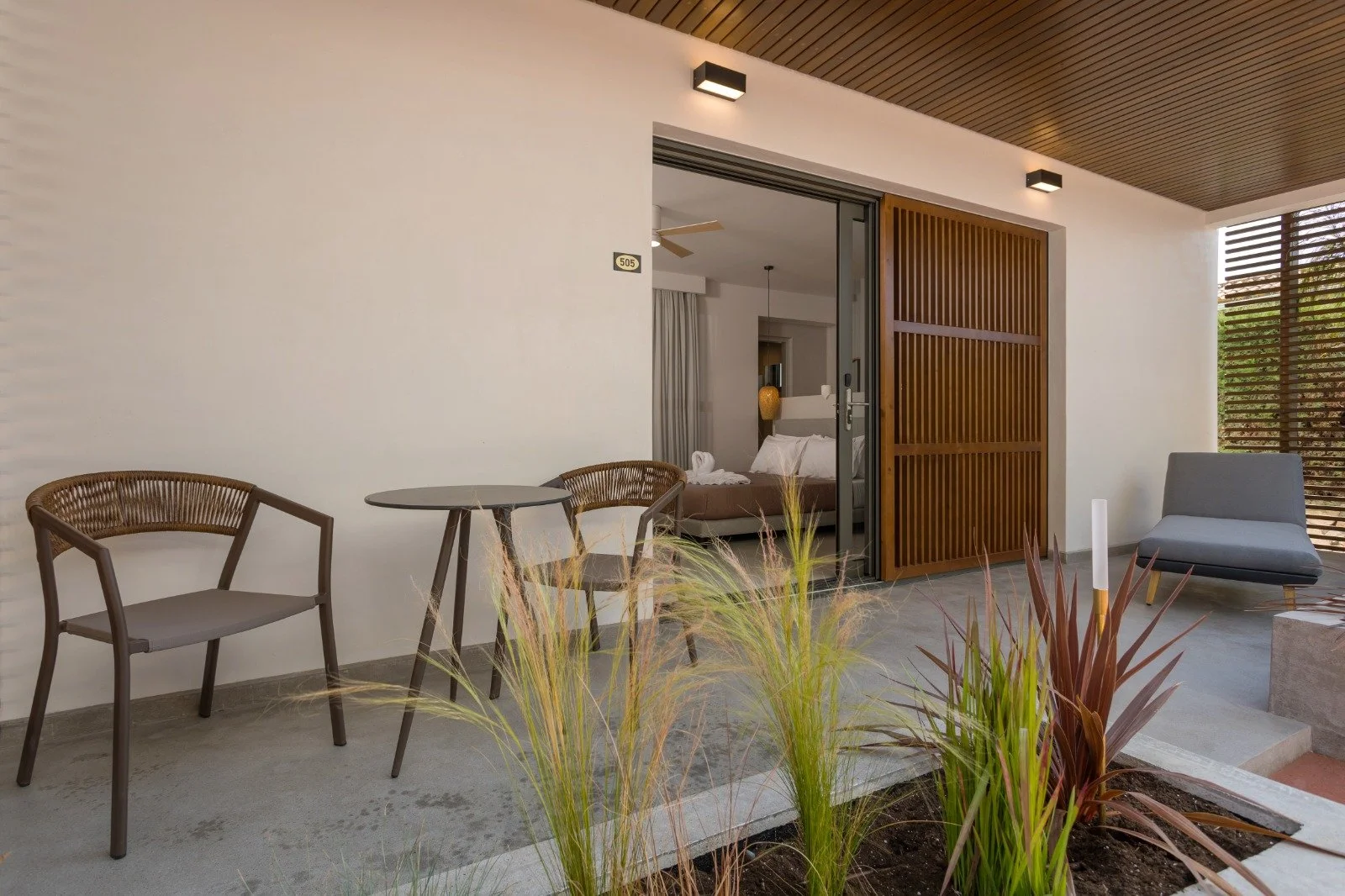 Modern hotel room balcony with outdoor seating, including a grey cushioned chair, a small round table, two brown chairs, and decorative plants. The balcony has wooden slats and sliding glass doors leading inside the room.