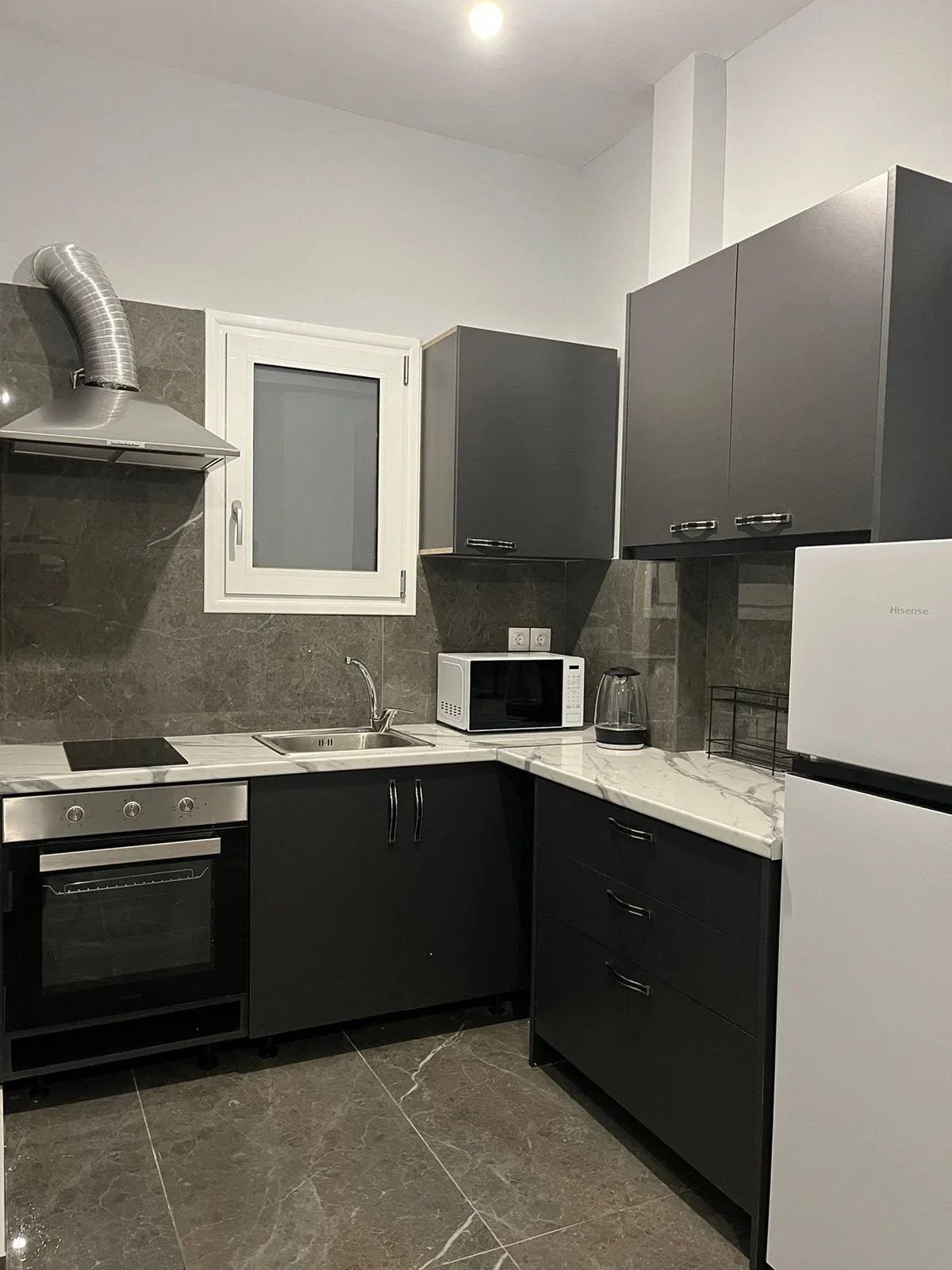 Modern kitchen with black and gray cabinets, marble countertops, and a stainless steel oven. Small window above the sink, microwave, coffee maker, and refrigerator are visible.