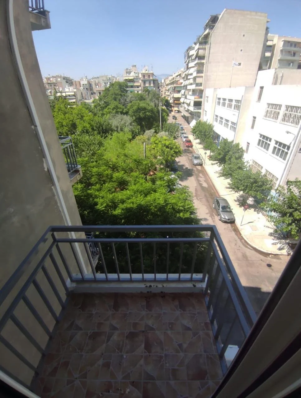 View from a balcony overlooking a city street lined with multi-story apartment buildings and green trees, with parked cars along the curb.