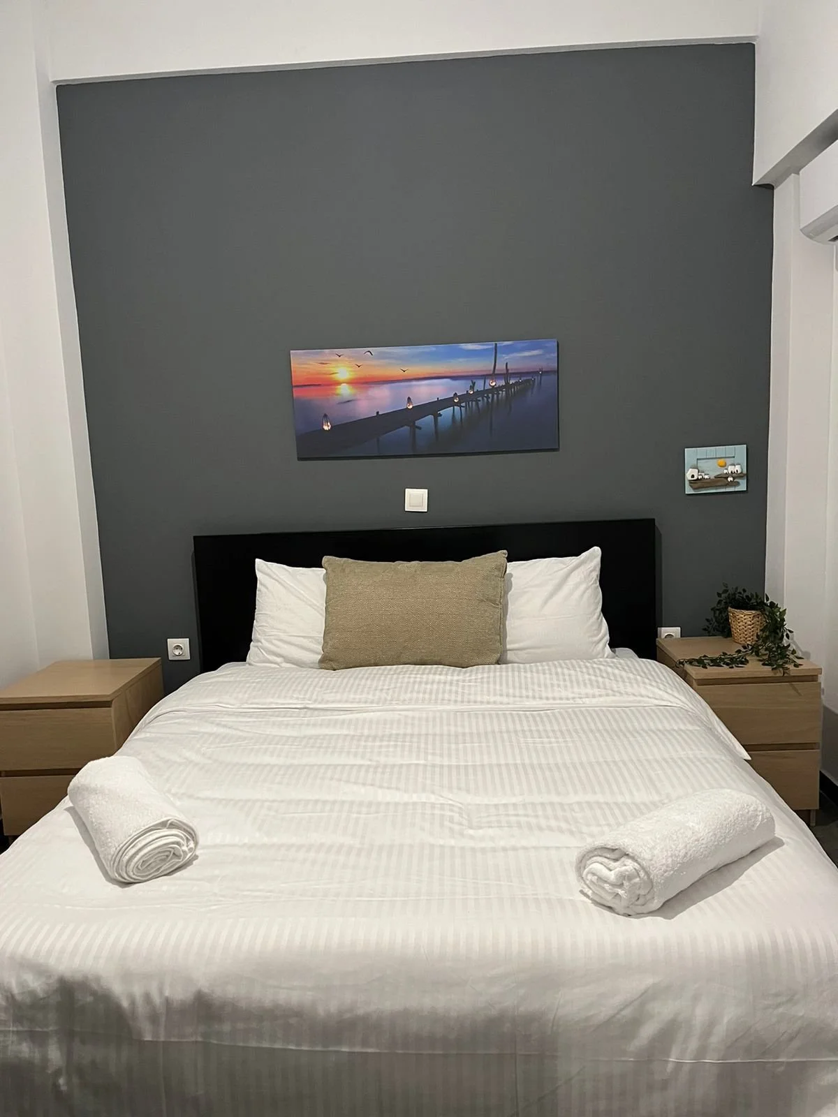 A neatly made bed with white linens and a beige pillow, flanked by two wooden nightstands, with two rolled towels on the bed. Above the bed, there is a dark gray accent wall with two landscape paintings, one of a sunset over water and a pier, and the
