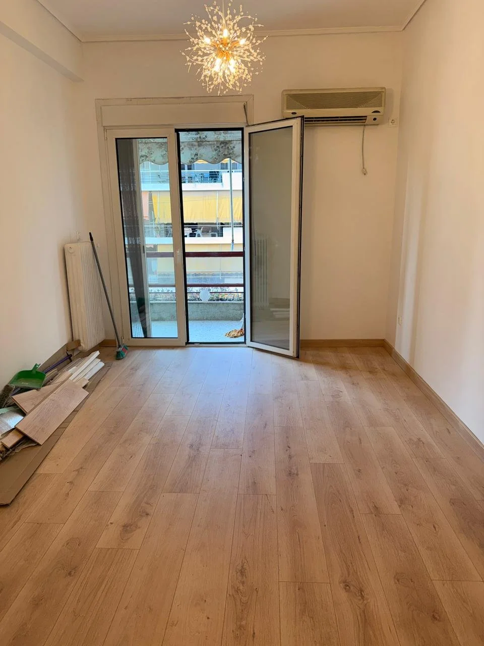 Empty room with wooden flooring, partially open glass door leading to a balcony, air conditioning unit above the door, wall socket, and cleaning tools in the corner.
