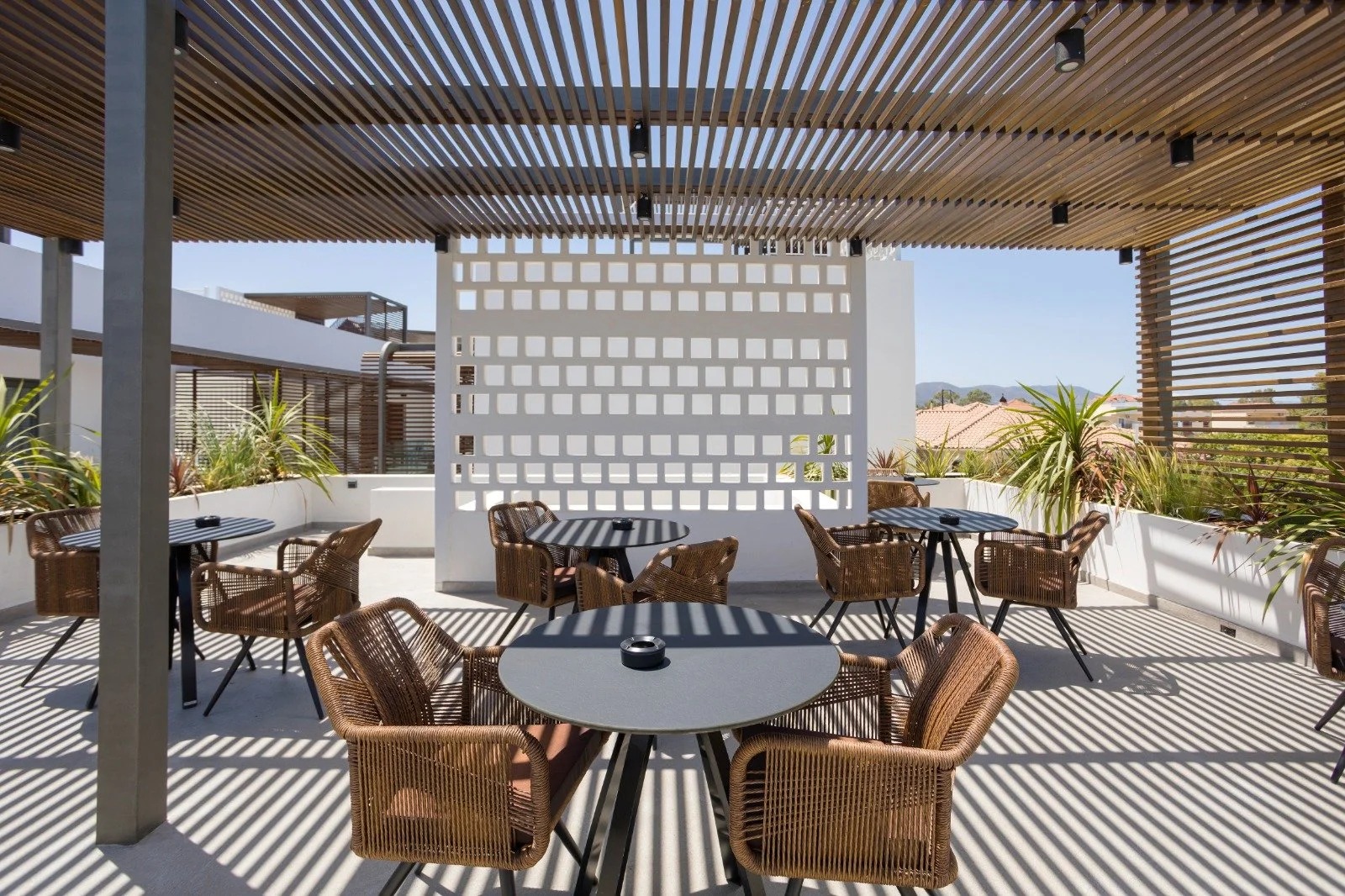 Outdoor rooftop patio with wicker chairs and round tables, plants in white planters, wooden slats casting striped shadows, and a partial view of the cityscape with rooftops and mountains in the distance.