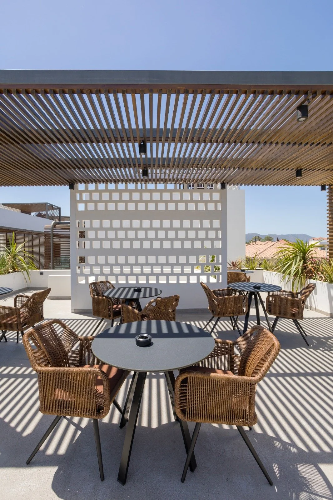 Outdoor rooftop patio with round tables and wicker chairs, with a wooden slat ceiling and sun shadows, against a backdrop of mountains and rooftops.