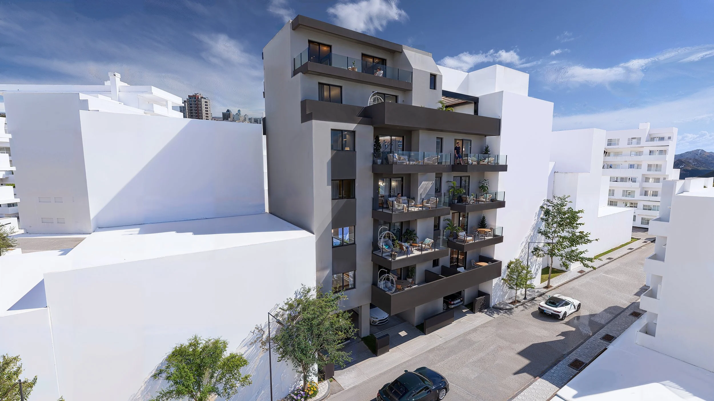 Modern multi-story apartment building with balconies, some with outdoor furniture and plants, situated along a street with parked cars and trees, under a partly cloudy sky.