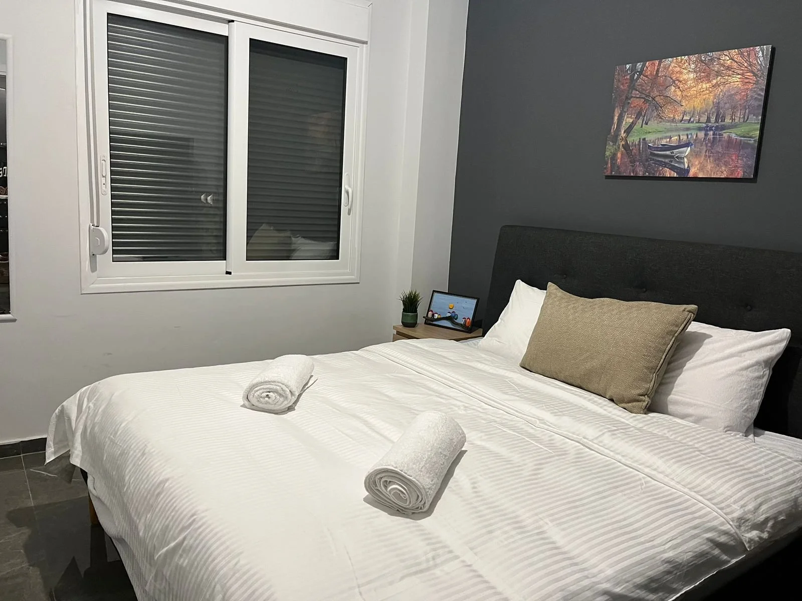A neatly made bed with white linens and two rolled towels on top, in a modern bedroom with dark gray accent wall, a window with blinds, and a framed landscape photo of a river and trees in autumn.