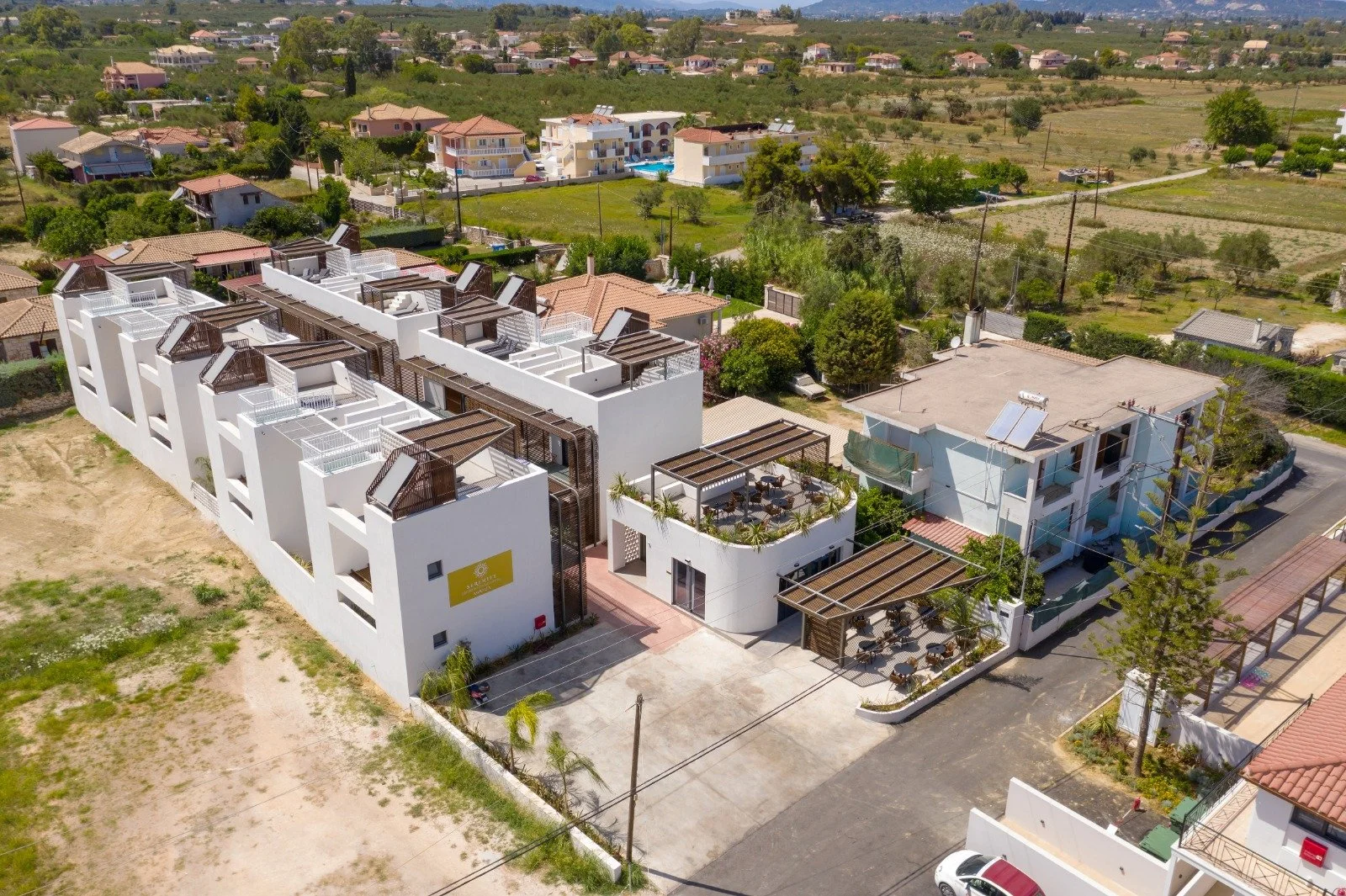 Aerial view of modern residential buildings with white facades and rooftop terraces, surrounded by a mix of greenery, trees, and neighboring houses in a suburban area.