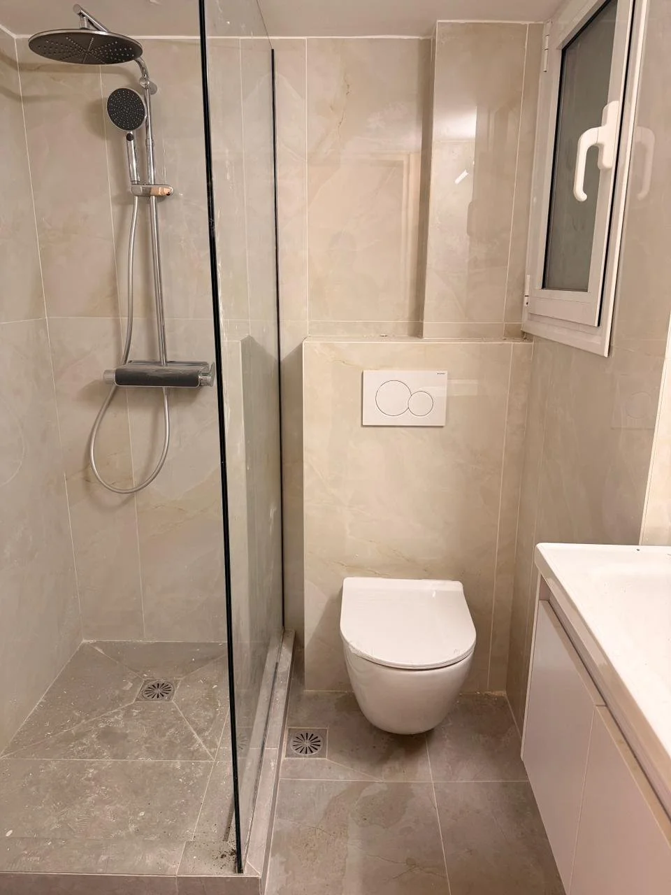 Bathroom with a walk-in shower, wall-mounted toilet, small window, and a white vanity cabinet.