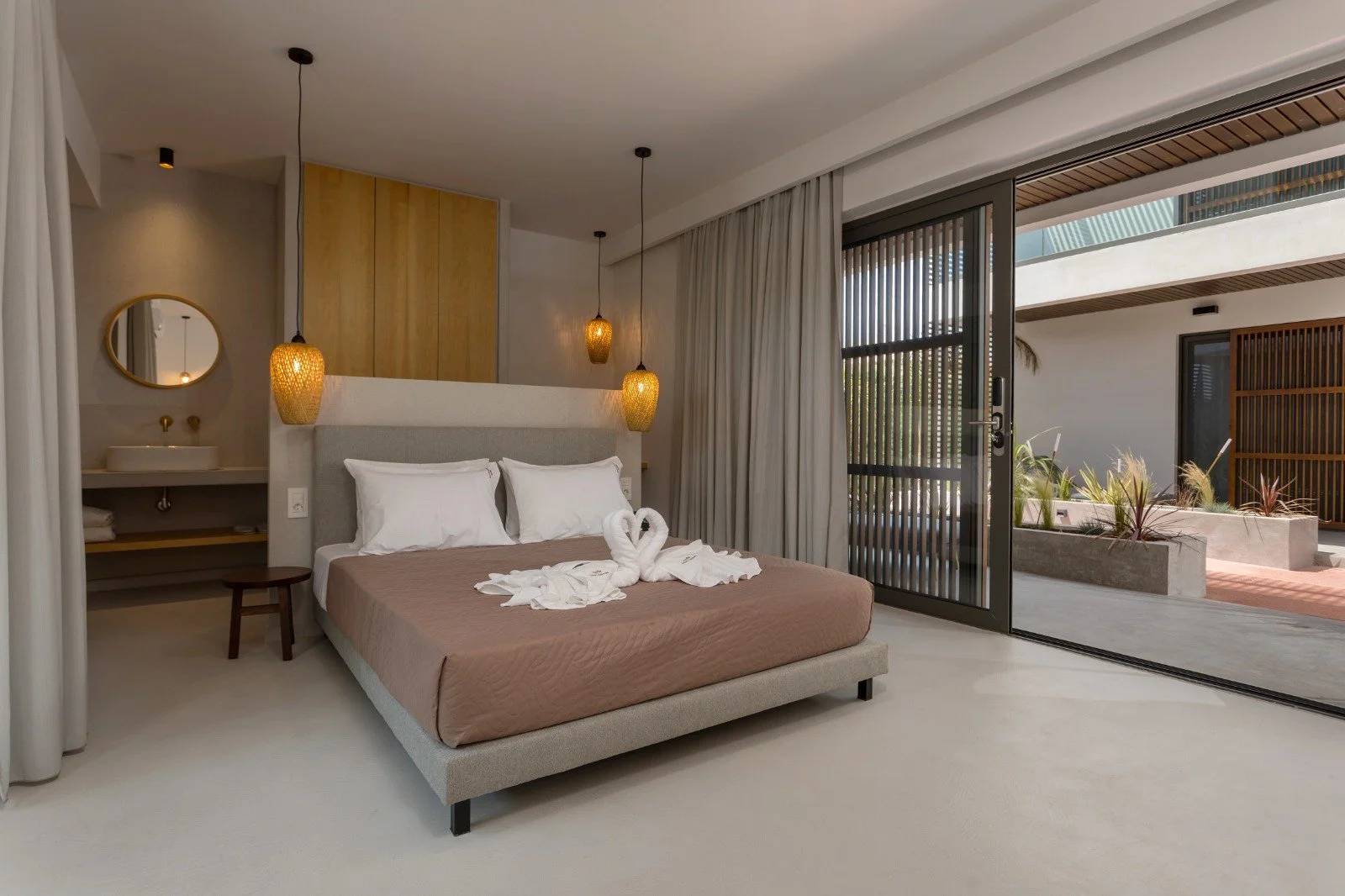 Modern bedroom with neutral tones, hanging pendant lights, a bed with white pillows, swan-shaped towels, and large sliding glass door leading to an outdoor patio with plants.