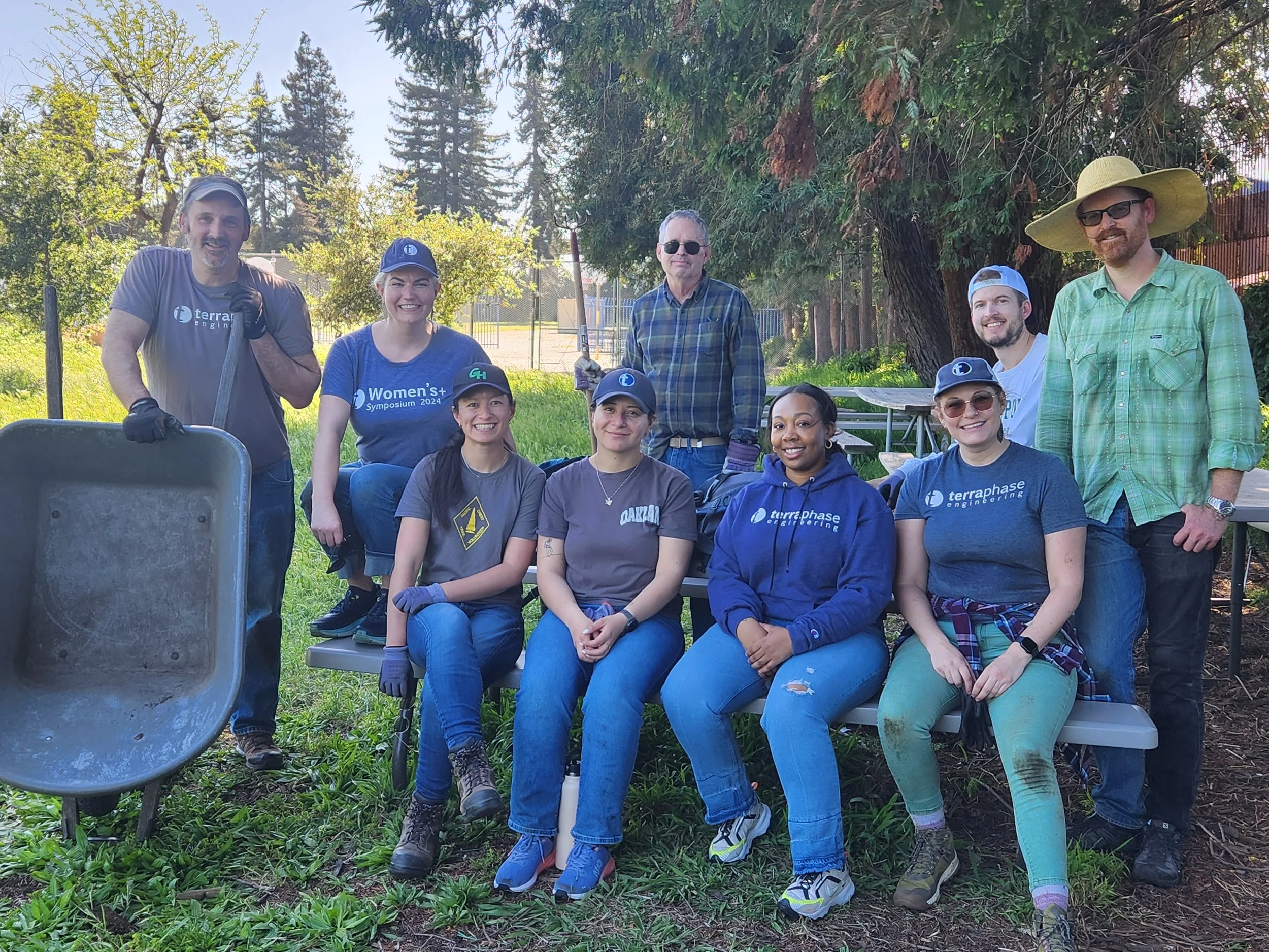 Group of ten people outdoors in a park, some wearing casual clothing and gloves, posing for a photo after a community service or volunteer event. They are smiling, and the background features trees and a sunny sky.