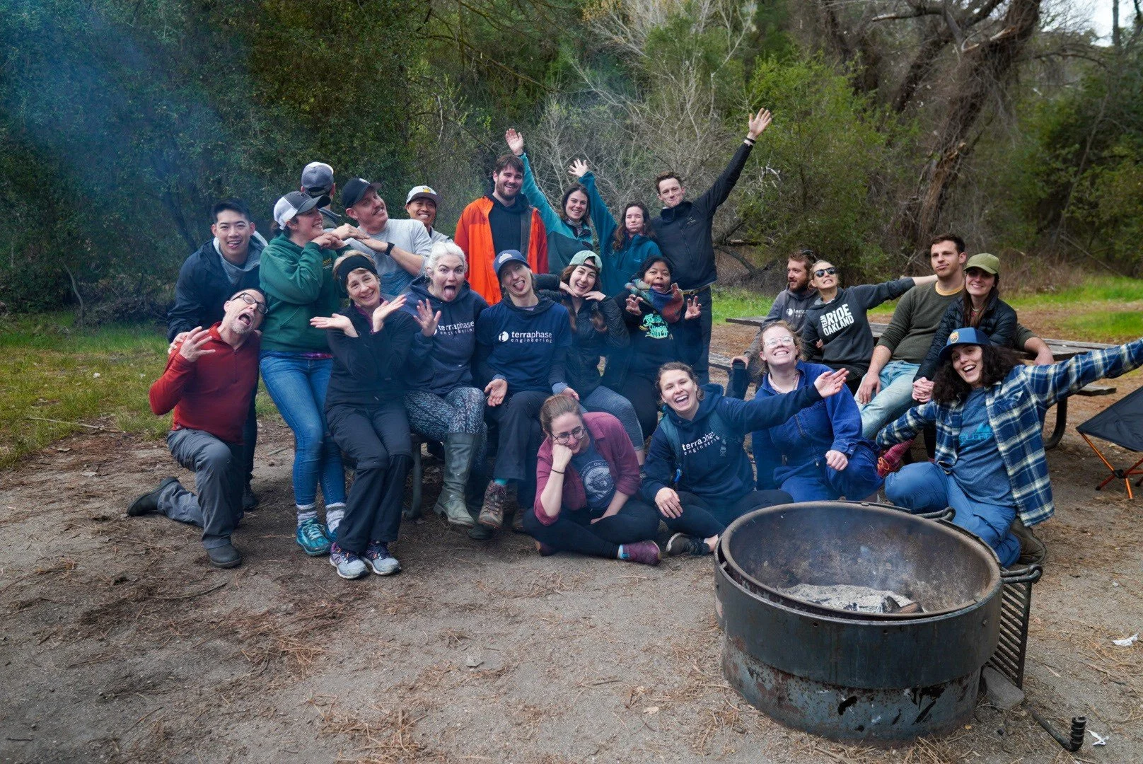 Group of people smiling, celebrating, and posing together outdoors near a campfire ring in a wooded area.