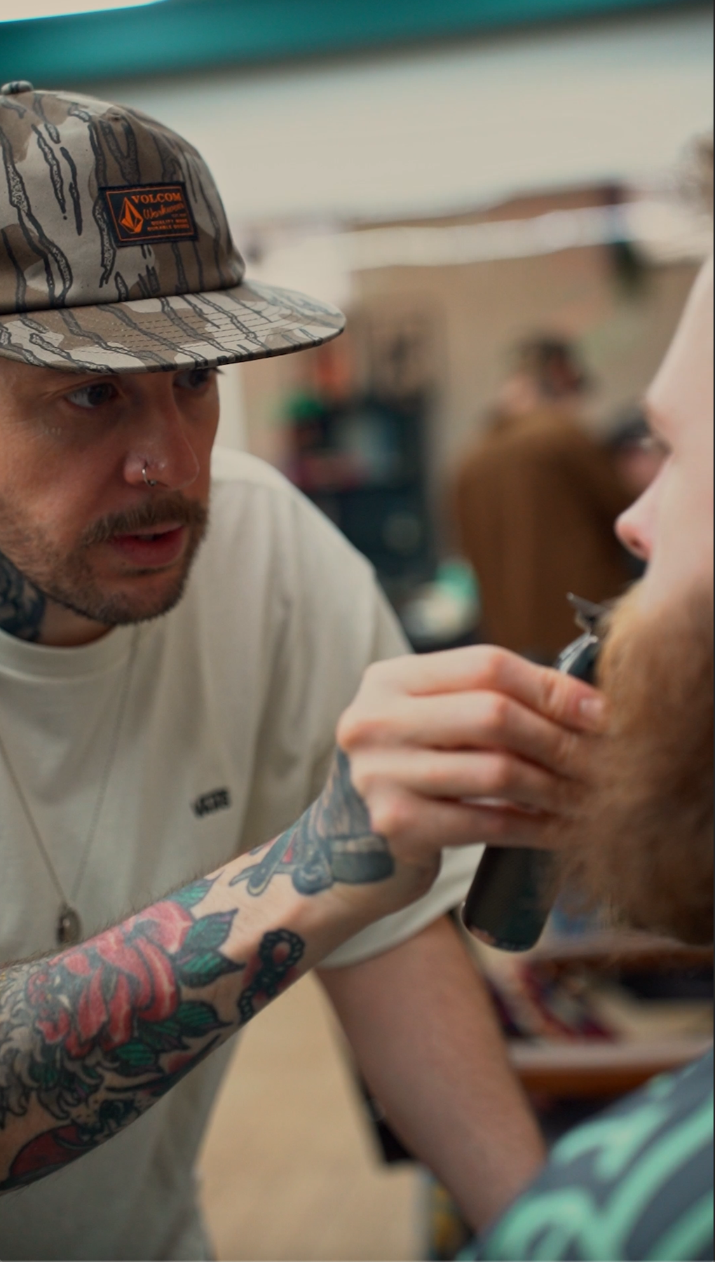 Tattooed man with a cap shaves another man's beard in a barbershop.