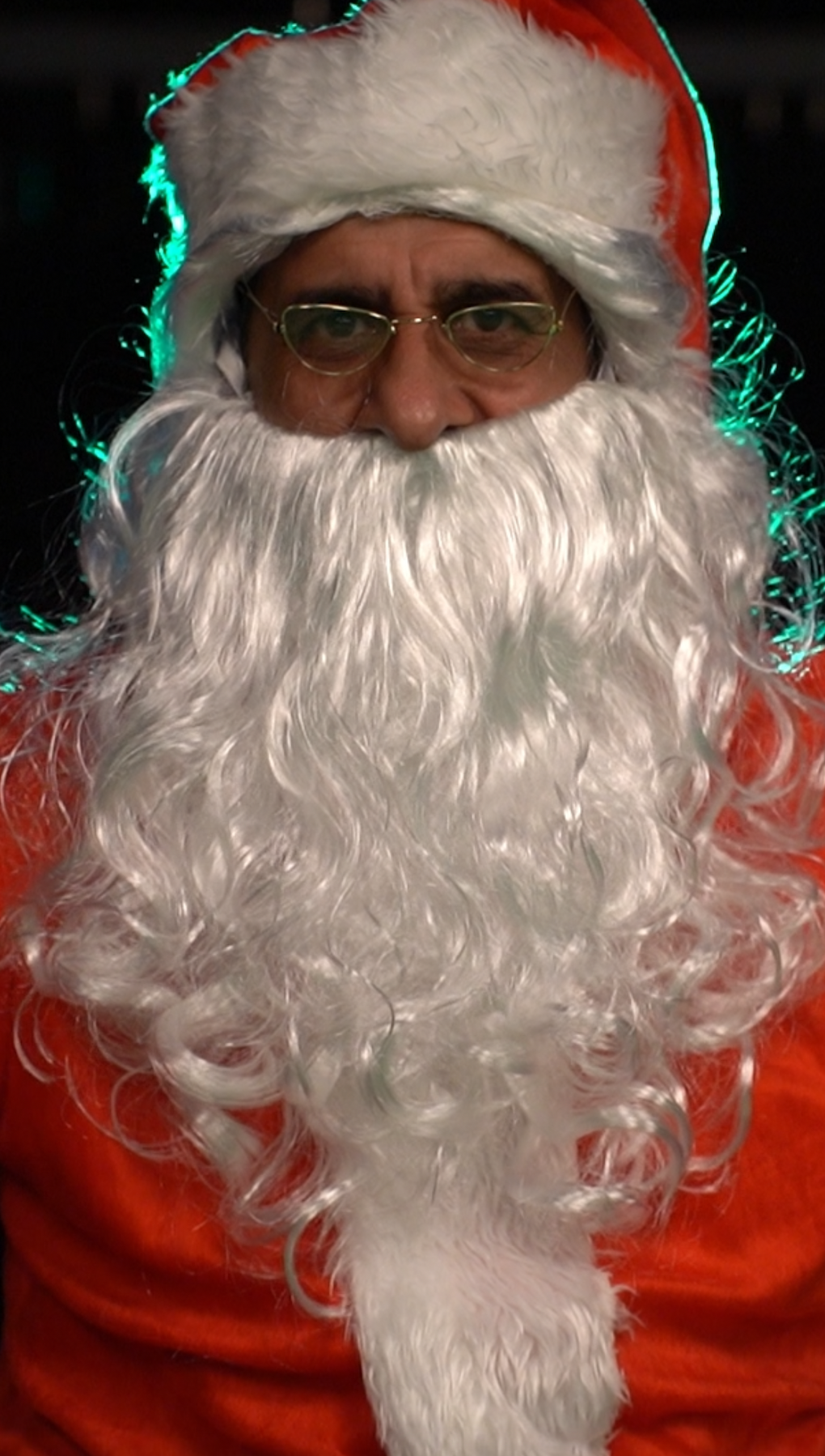 A man dressed as Santa Claus with glasses, a red suit, and a long curly white beard, taking a close-up photo.