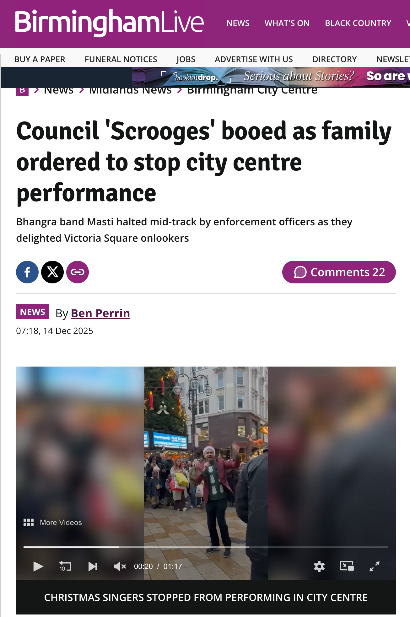 A person in a red jacket and Santa hat addressing a crowd around Christmas decorations and a Christmas tree in a busy city square, during the holiday season.