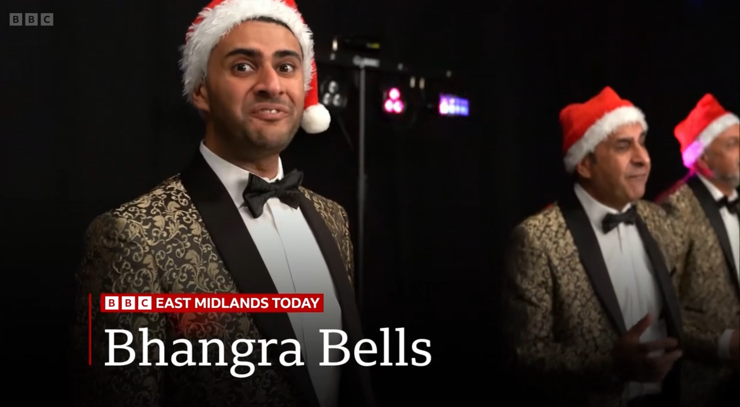 Three men in tuxedos with gold-embossed jackets and black bow ties, wearing red Santa hats, standing on a stage at a Christmas event. The background is dark with some colorful lights, and there is a