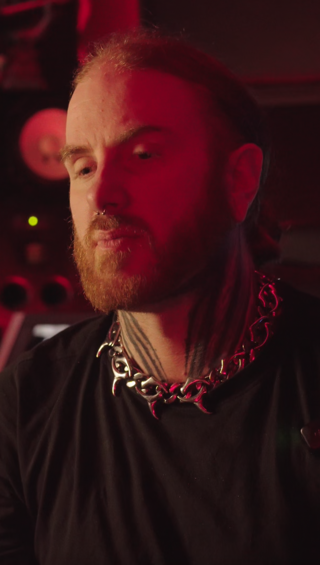 A person with long hair and a beard wearing a silver chain necklace and a black shirt in a dimly lit environment with red lighting.
