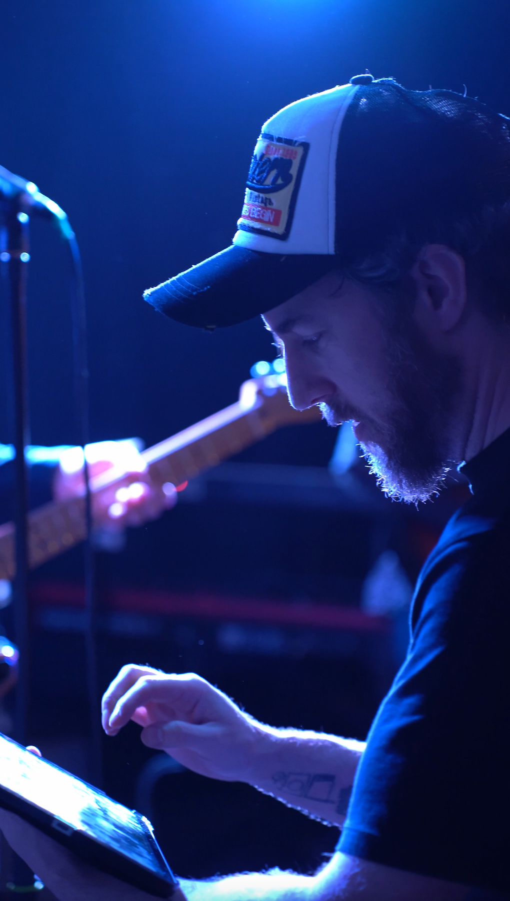 A man with a beard and a cap looking at a tablet in a dimly lit setting, with a guitar in the background.