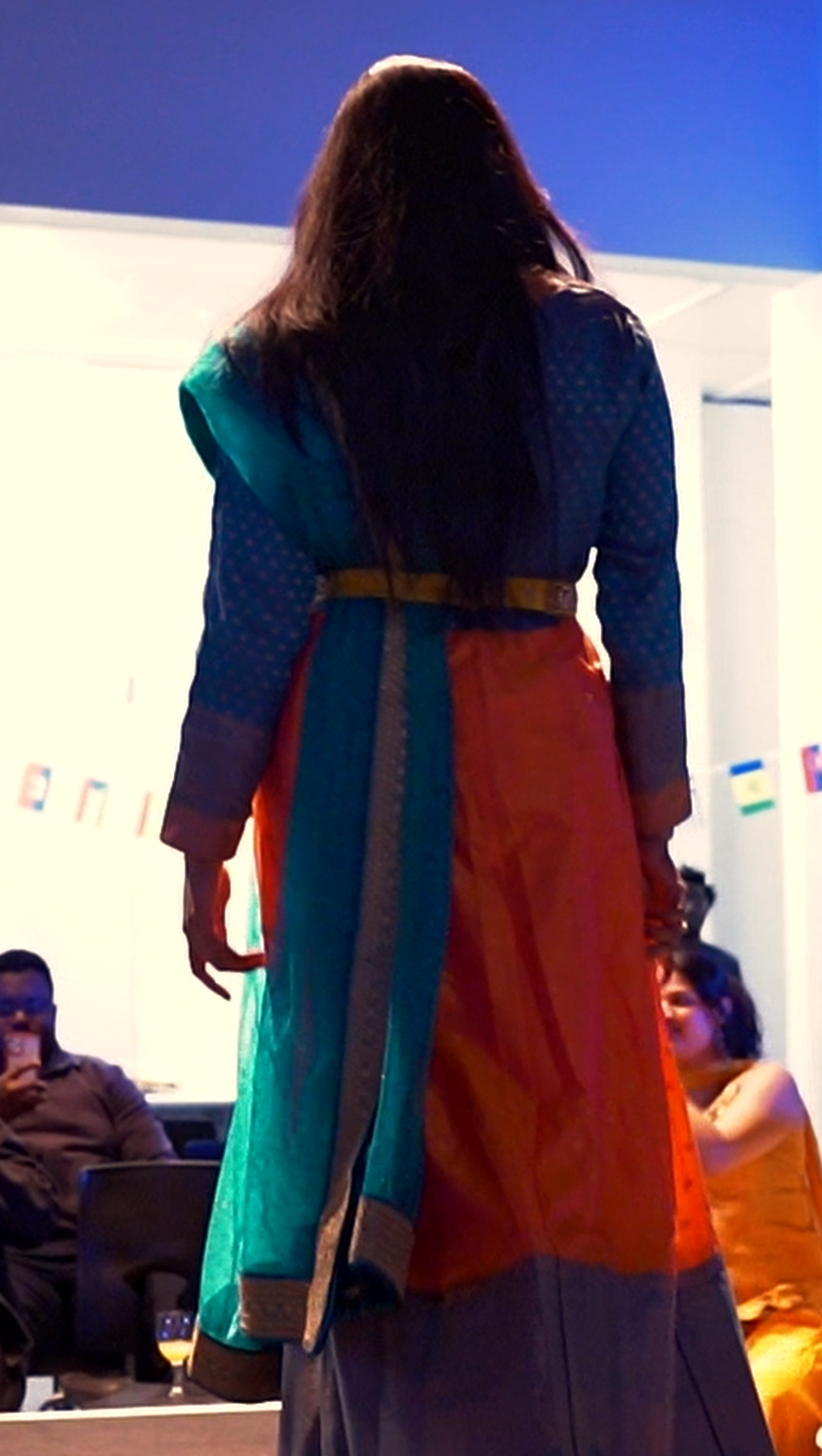 A woman with long dark hair seen from the back wearing colorful traditional Indian attire, including a blue and orange sari or dress, standing in a room with people sitting and using phones.