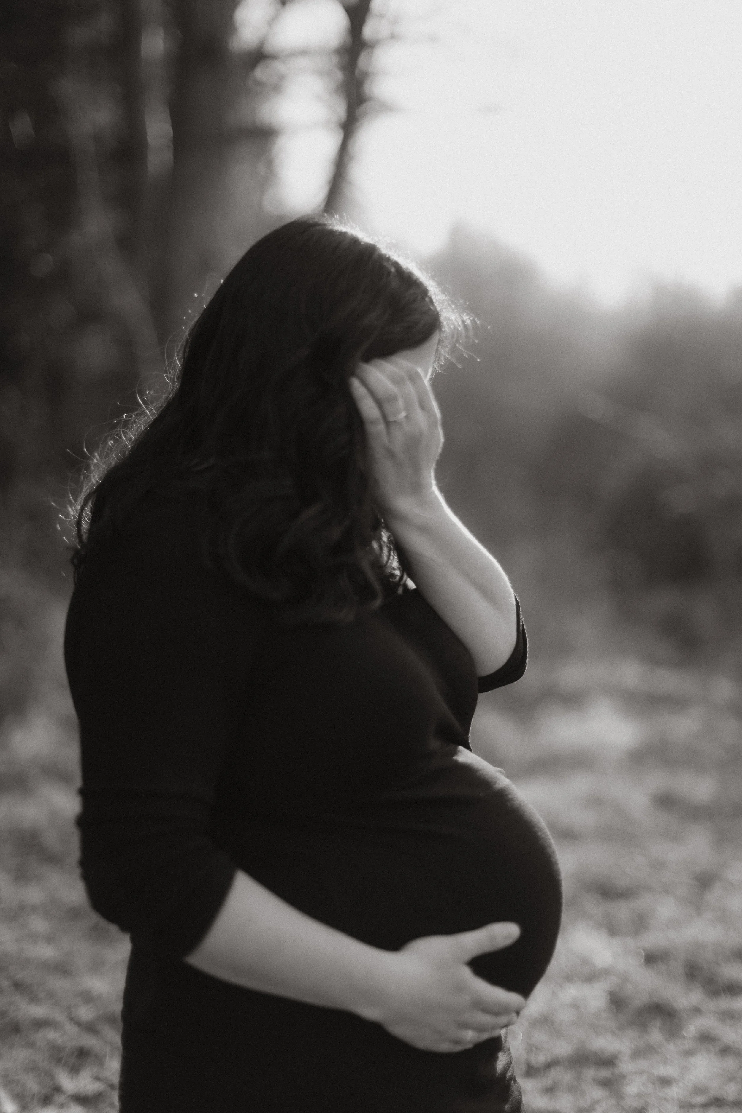 Schwangere Frau steht im Freien mit verschränkten Armen um den Bauch, in Schwarz-Weiß