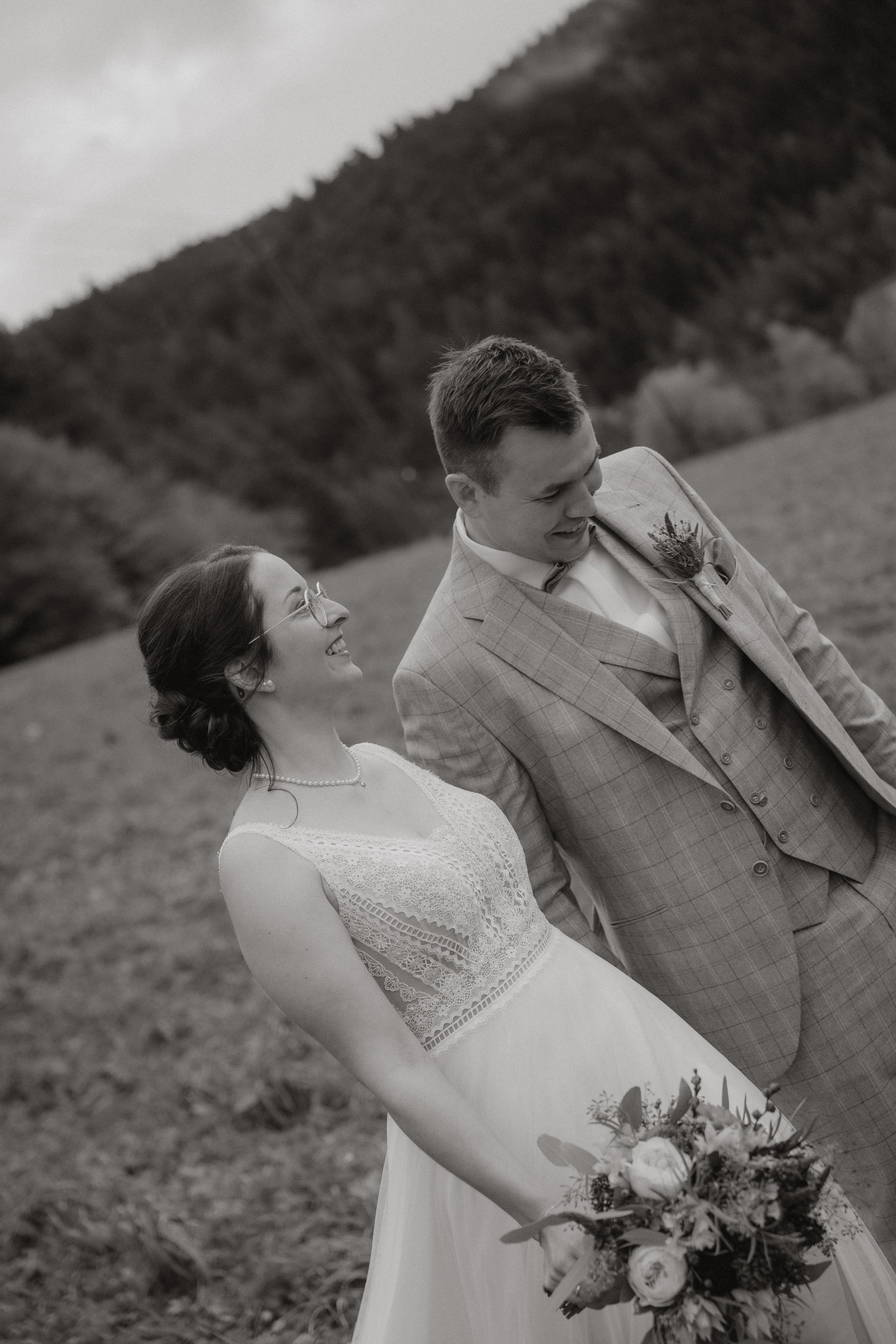 Schwarz-weiß-Foto eines Brautpaares, das im Freien auf einer Wiese mit Hügel im Hintergrund steht, beide lachen und schauen sich an, die Braut hält einen Blumenstrauß.