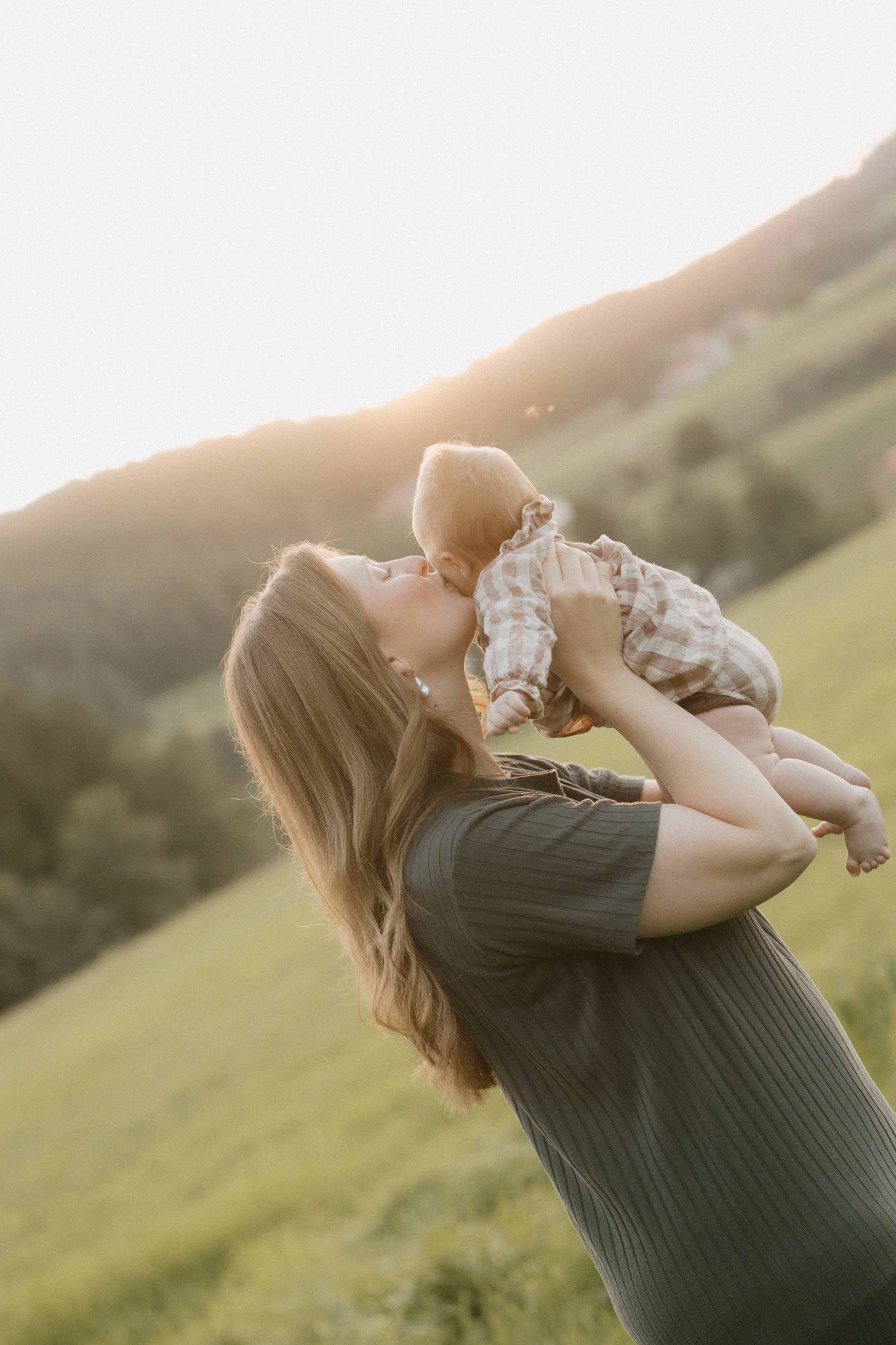 Eine Frau hält ein kleines Baby hoch in einer grünen, hügeligen Landschaft bei Sonnenuntergang und küsst es.
