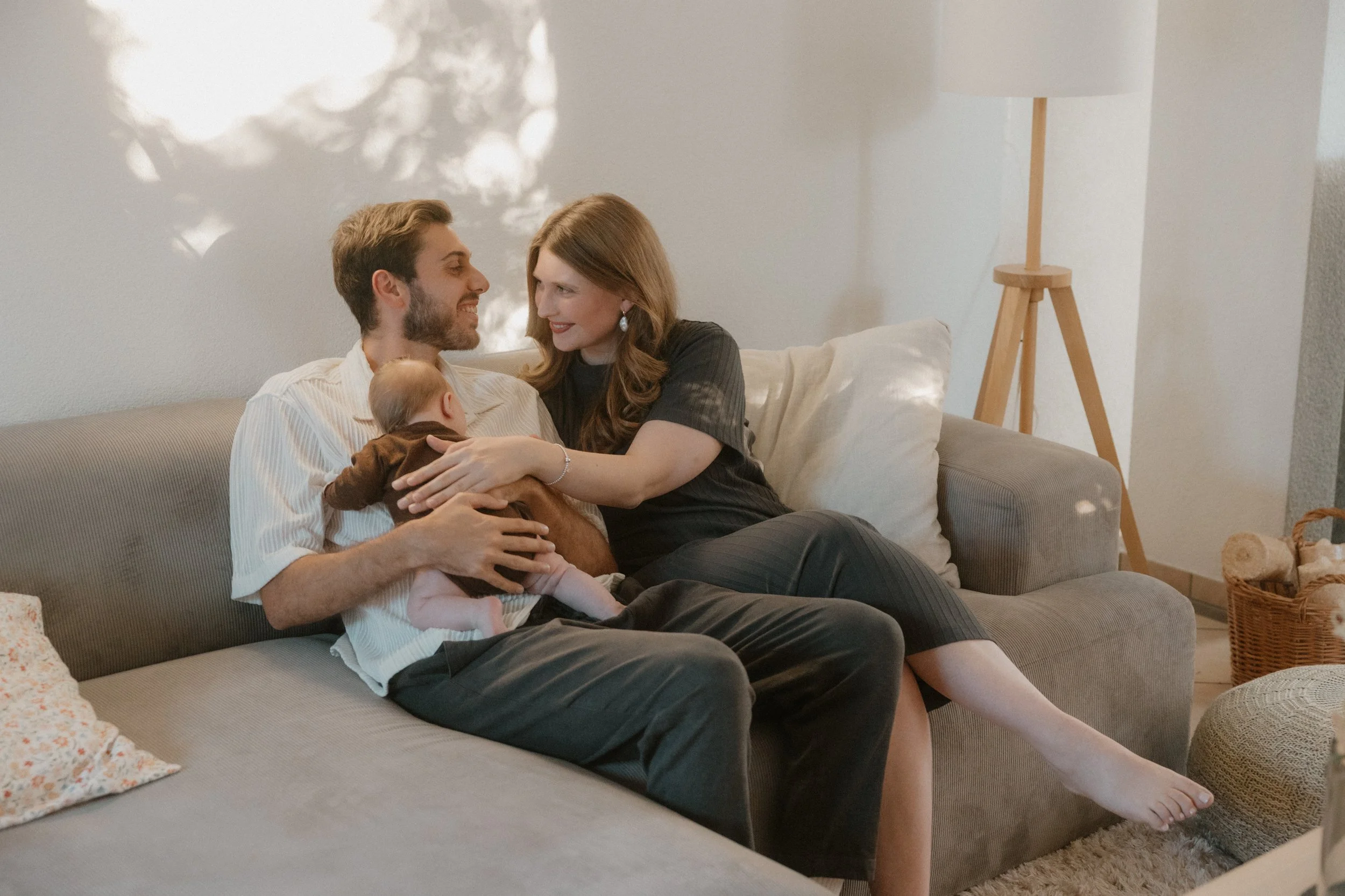 Ein Paar sitzt auf einem sofa und hält ihr Baby im Arm, sie lachen und schauen sich liebevoll an.