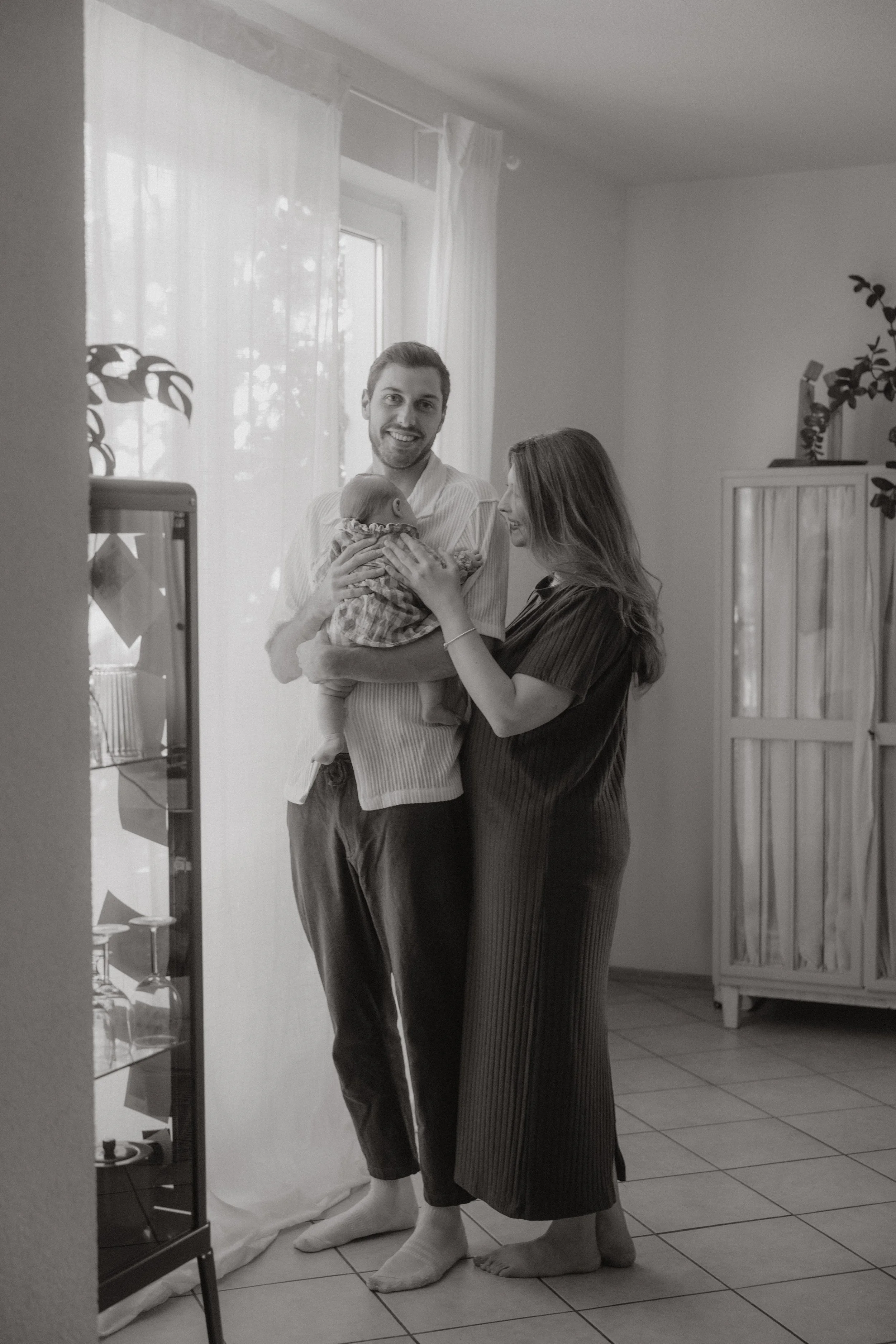 Ein glückliches Paar hält ein neugeborenes Baby in einem hellen Raum mit Gardinen und Pflanzen.