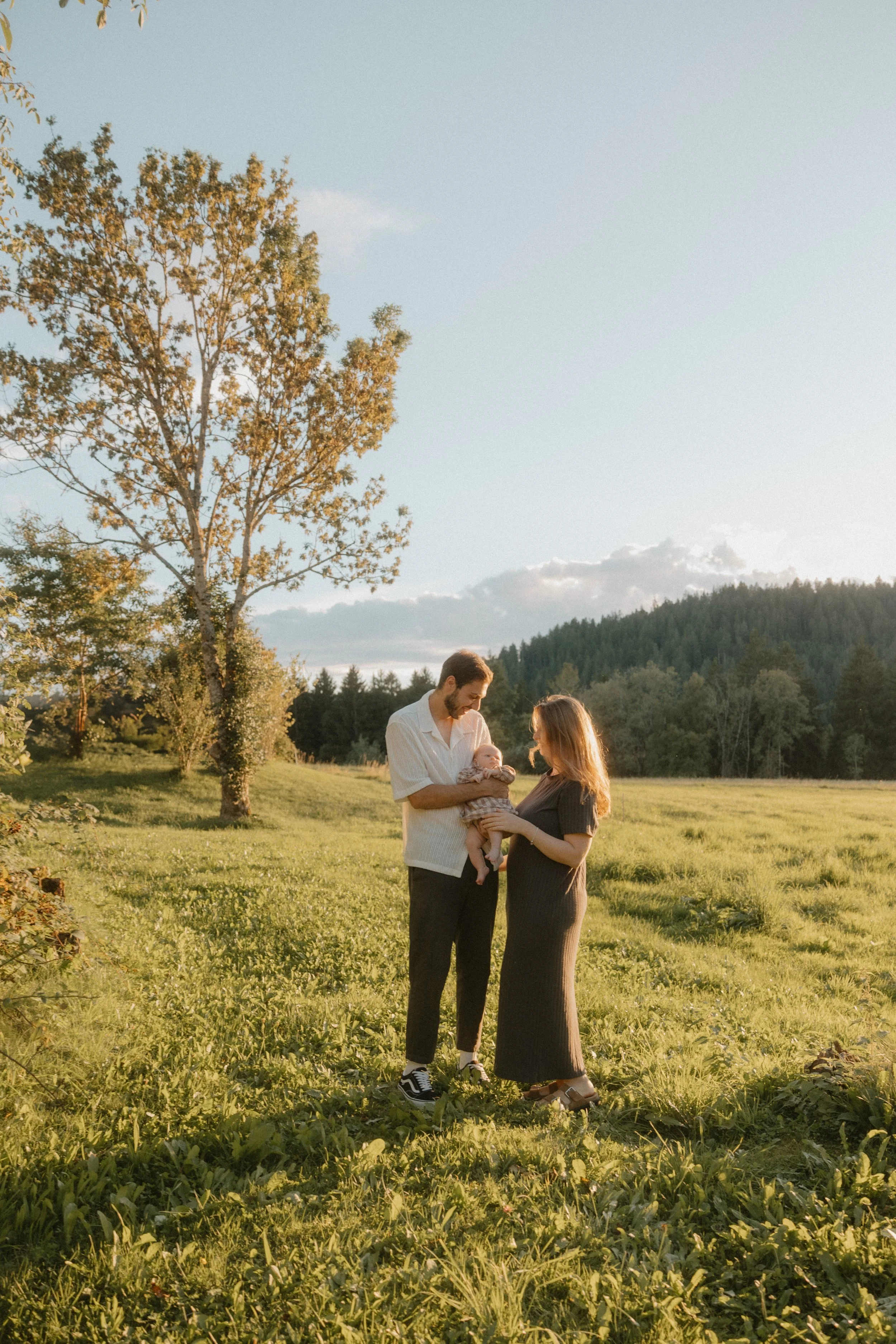 Ein Paar mit einem Baby im Freien bei Sonnenuntergang, umgeben von grünen Wiesen und Bäumen, in ländlicher Umgebung.
