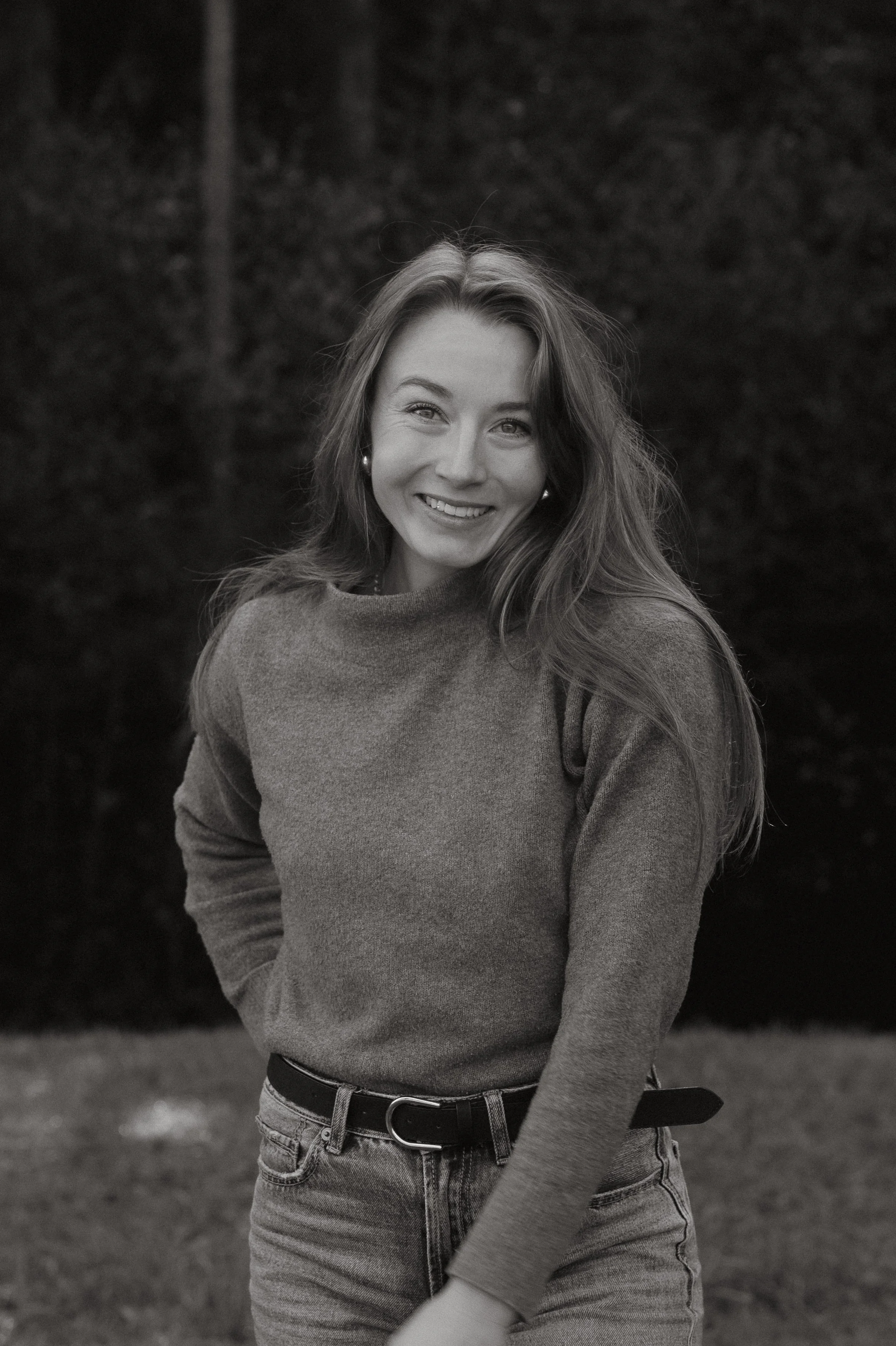 Schwarz-Weiß-Foto eines lachenden jungen Mädchens mit langem Haar, das draußen auf einer Wiese steht, wearing a sweater und Jeans.