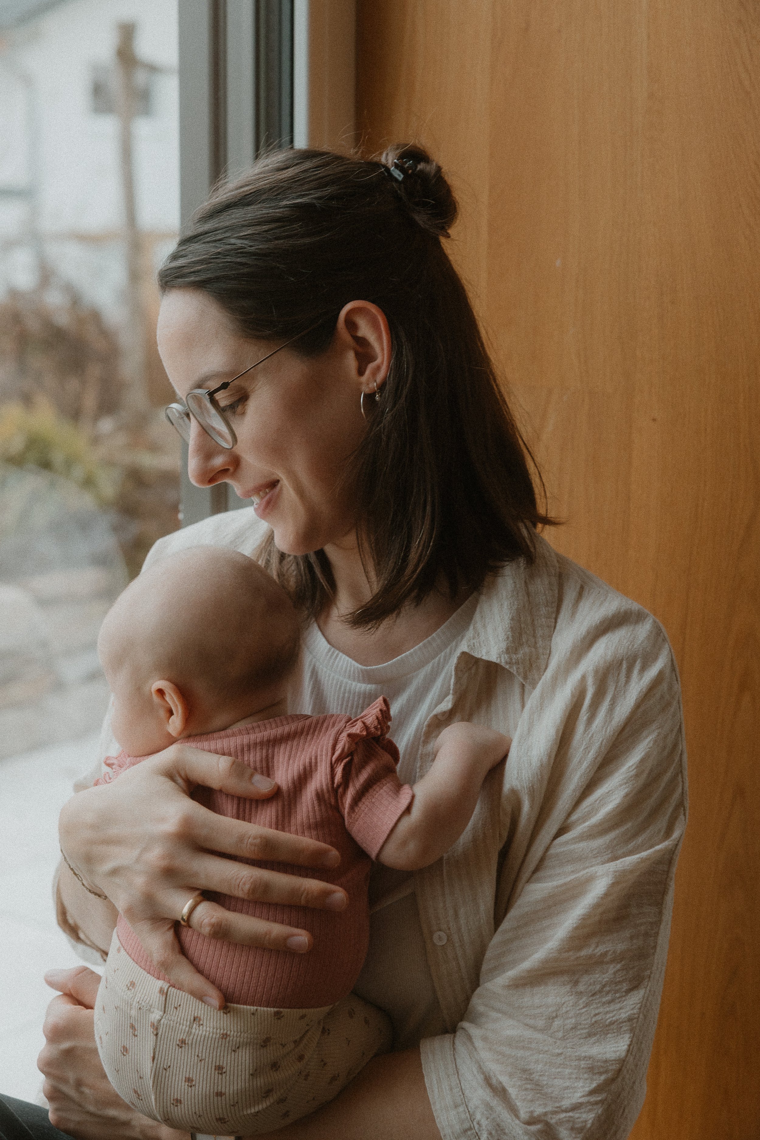 Eine Frau hält ein Baby, das in ihren Armen liegt, vor einem Fenster, lächelt und schaut liebevoll auf das Baby, während es an ihrer Schulter schnuppert.