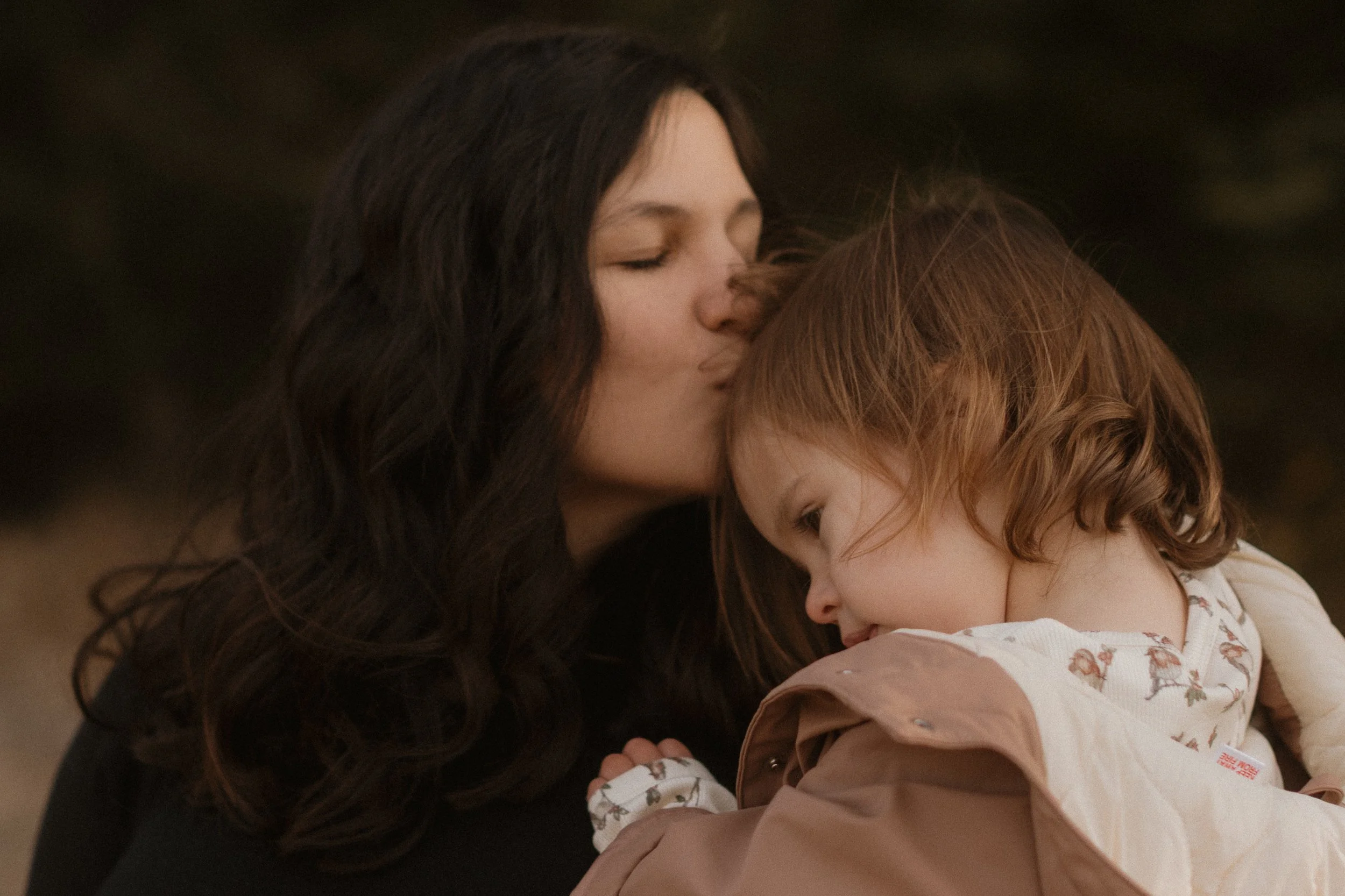 Eine Frau mit schwarzen, lockigen Haaren küsst ein schlafendes kleines Mädchen mit roten Haaren auf den Kopf.