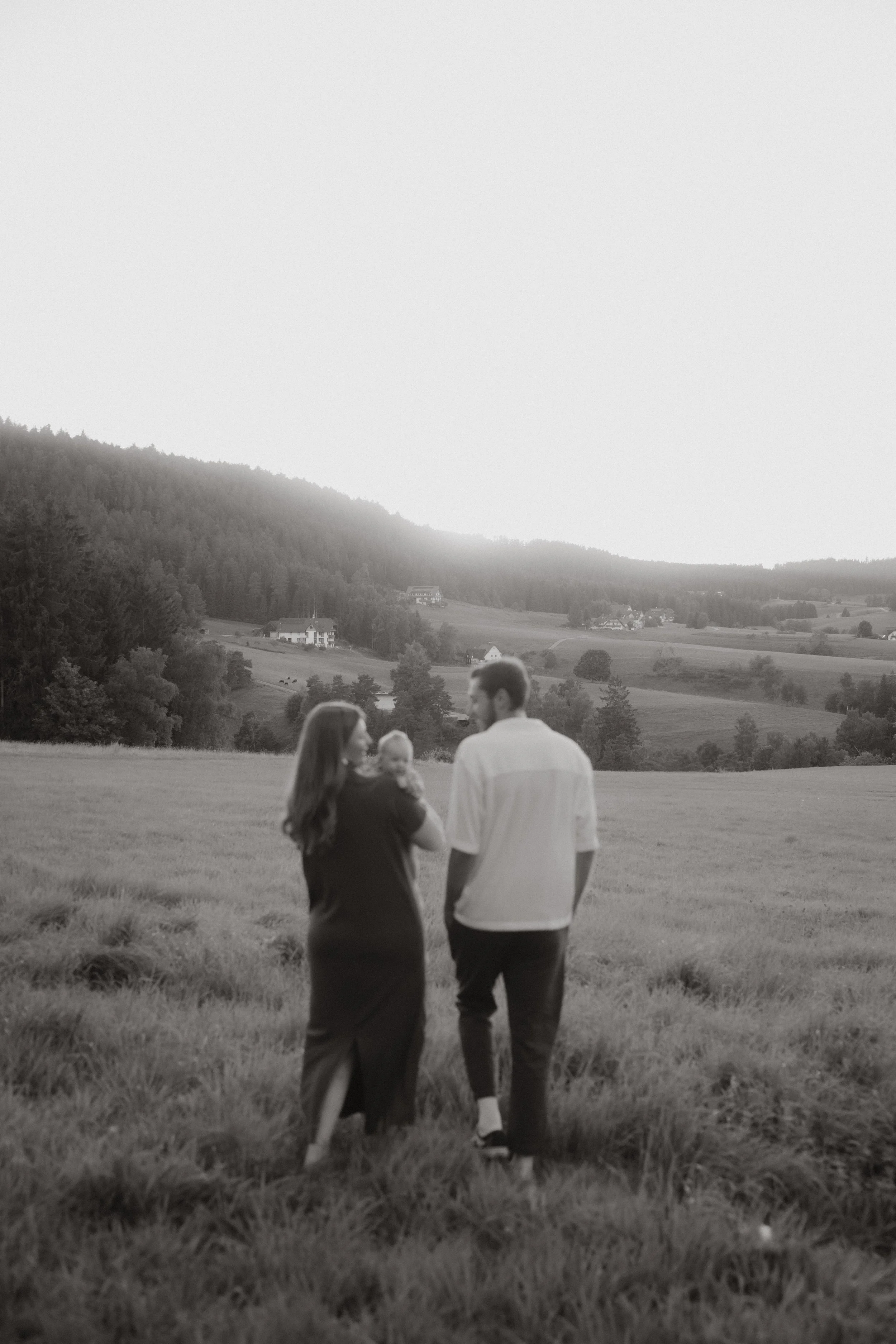 Ein Paar geht mit einem Baby in einem offenen Feld, umgeben von Hügeln und Bäumen, bei Sonnenuntergang, schwarz-weiß