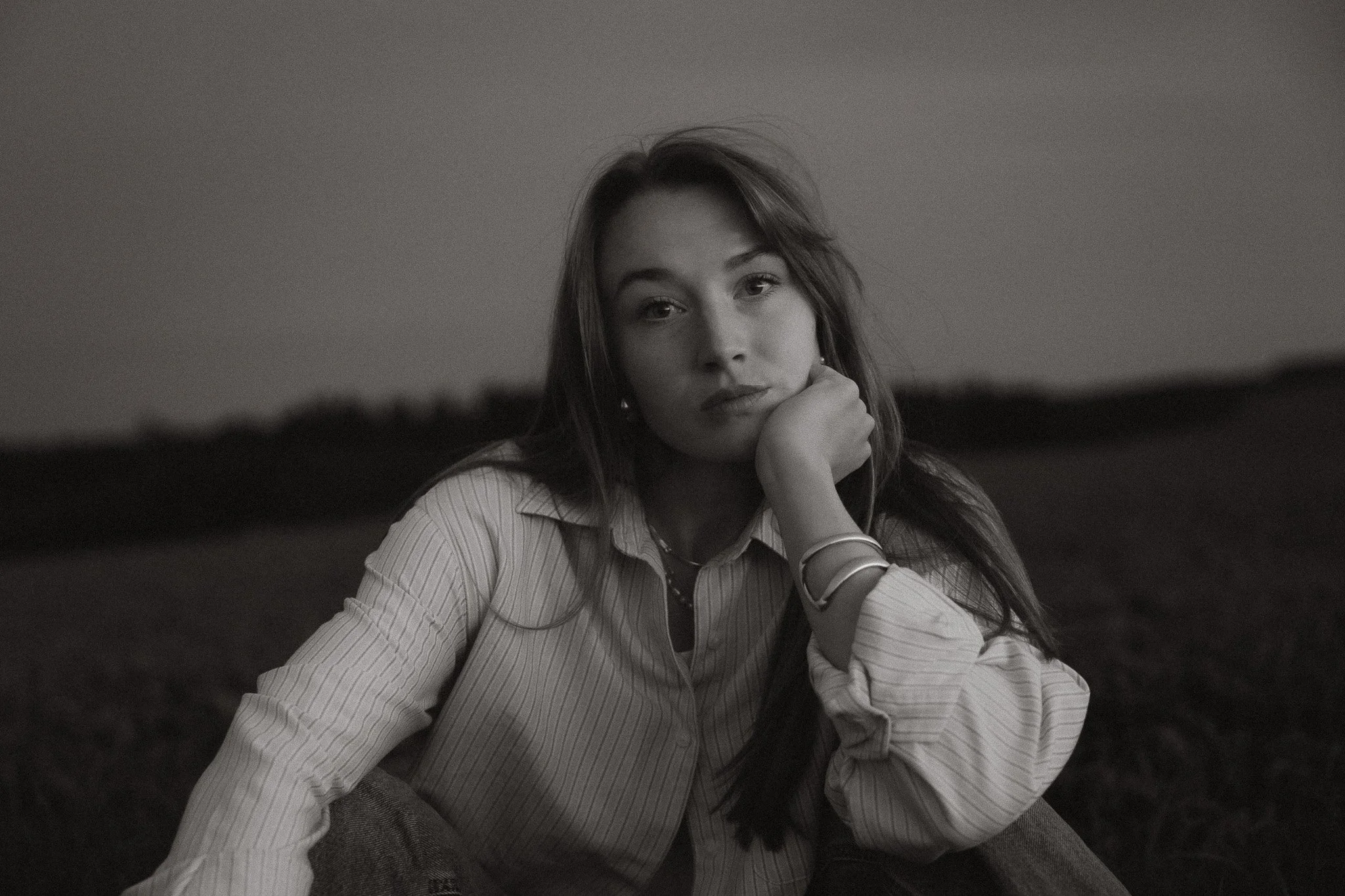 Schwarz-Weiß-Foto eines nachdenklichen jungen Mädchens, das im Freien sitzt, mit lockigem Haar, trägt eine gestreifte Bluse und mehrere Armbänder, Blick direkt zur Kamera, im Hintergrund unscharfe Natur.