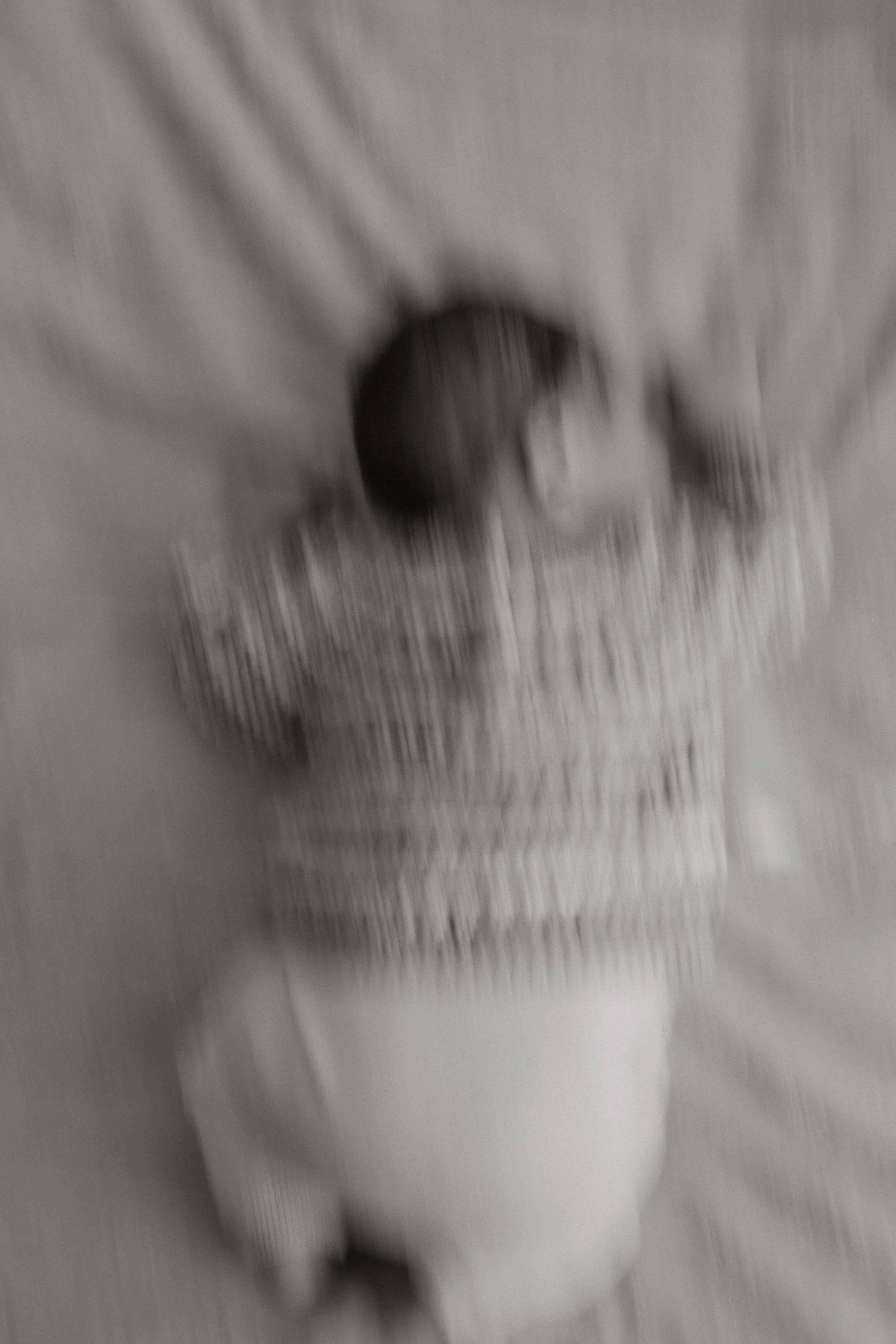 Ein Baby liegt auf einer weichen Oberfläche, vermutlich eine Decke, und trägt ein gestricktes Kleid. Das Bild ist unscharf und in Schwarz-Weiß.